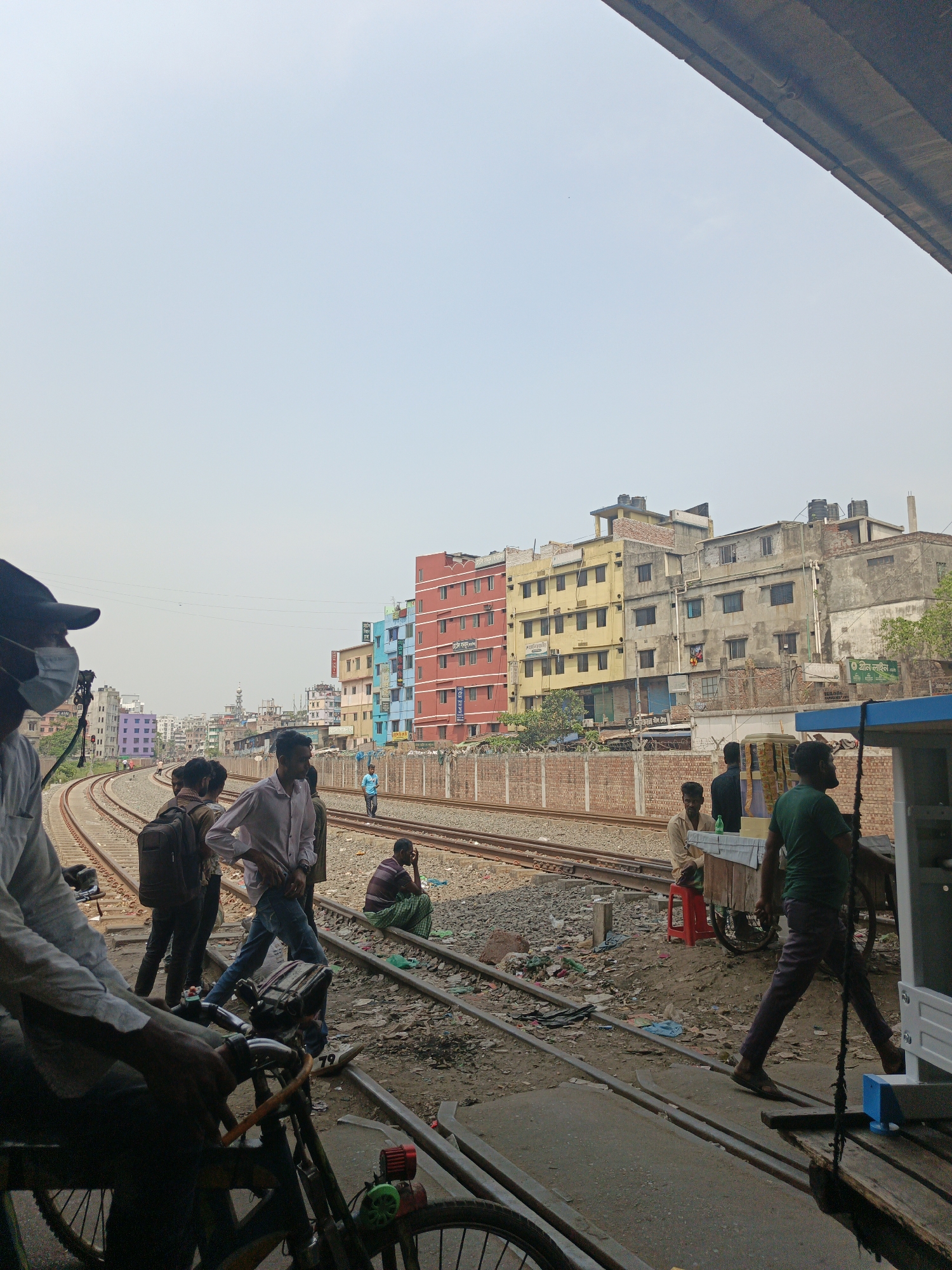 The image shows a scene in Dhaka, Bangladesh, near railroad tracks. Several people are present, some standing or walking on the tracks, others sitting or waiting. A person on a bicycle is in the foreground. Buildings, a brick wall, and some trash and debris are visible along the tracks. There is a food cart vendor in the scene. The sky is overcast.