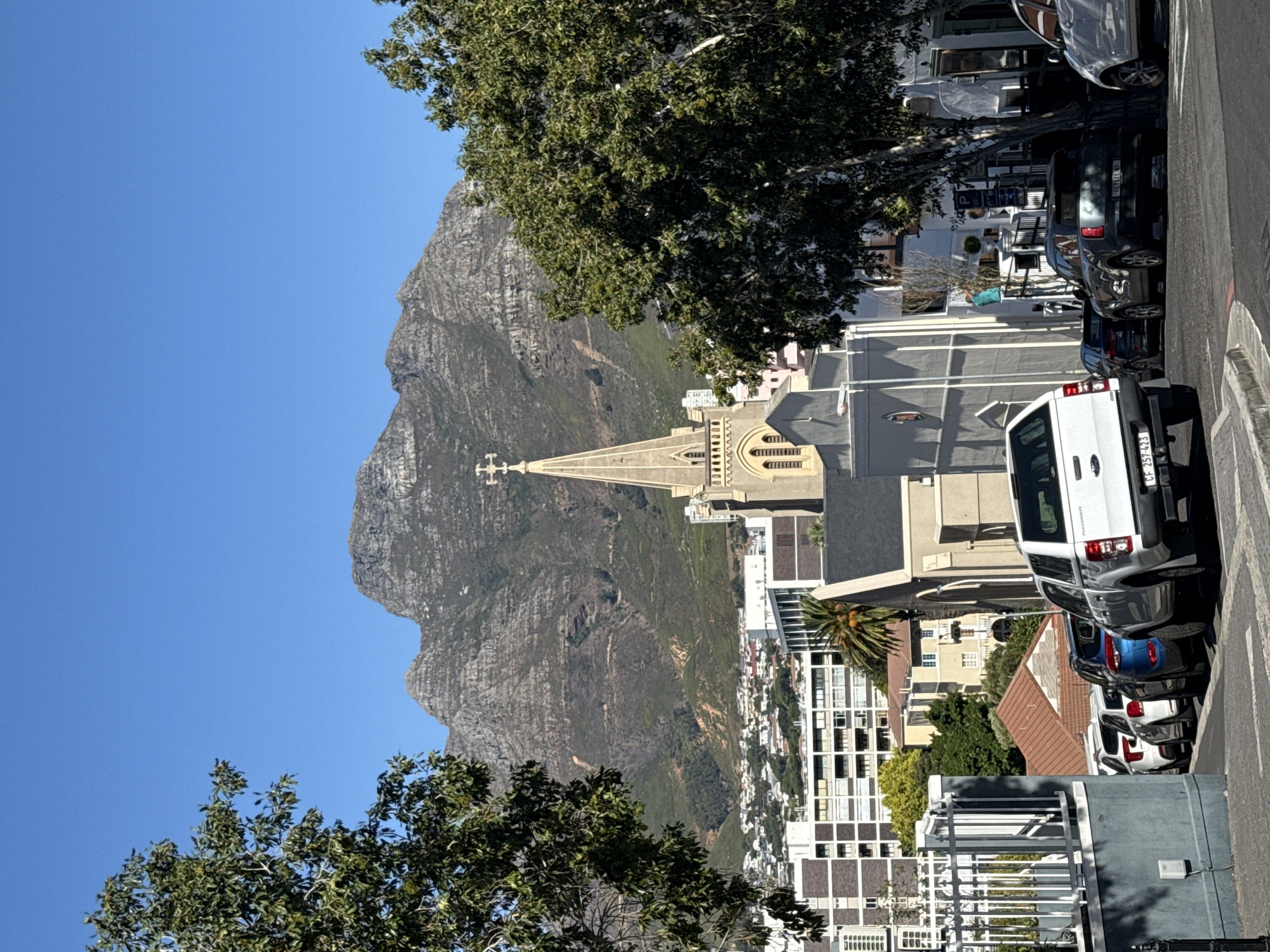 The image depicts a street scene in Cape Town, South Africa, with Table Mountain in the background. A church with a tall steeple and cross is prominently featured. Several parked cars line the side of the street. Buildings of various heights are also visible, some with palm trees nearby. A tree with green foliage is in the foreground on the left. The sky is clear and blue.