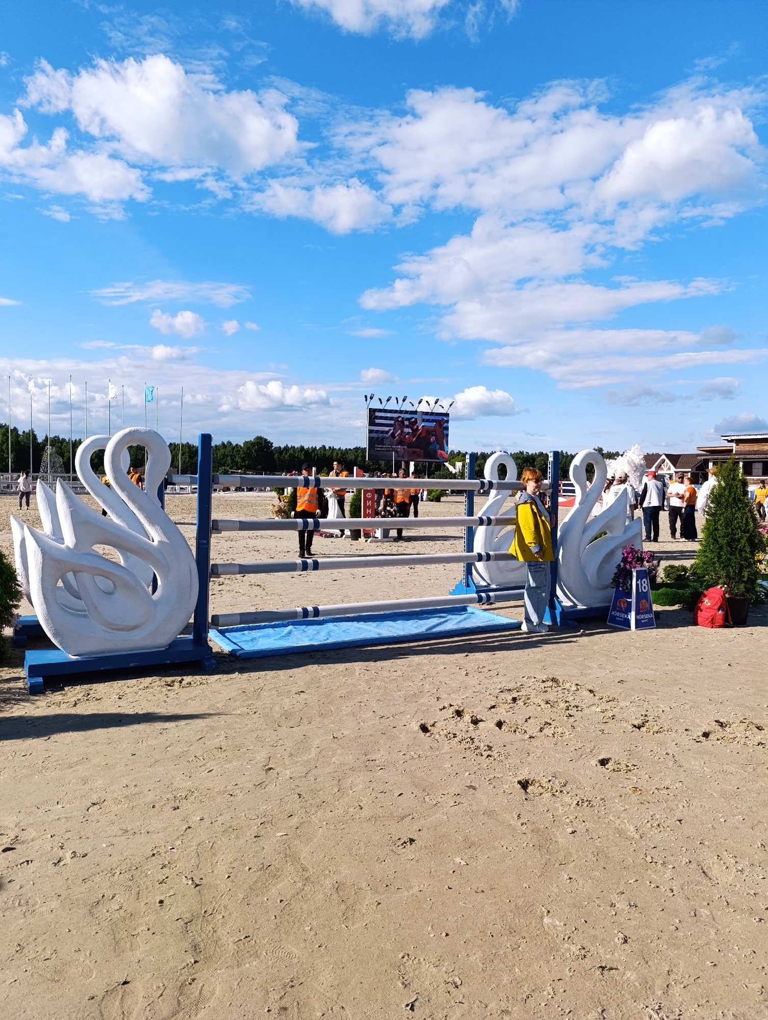 The image depicts an outdoor equestrian event, likely a show jumping competition, set on a sandy arena. The foreground features a decorative obstacle with two large, white swan sculptures as its base. The swans are positioned on blue platforms. In the mid-ground, several horizontal poles, also blue and white, form part of the jump. A young person wearing a yellow jacket and jeans stands near the swan sculptures, facing the camera. Behind the jump, a crowd of people is visible, some wearing orange vests, suggesting they are event staff. Further in the background, trees line the perimeter of the arena, and a large screen displays images, possibly of the ongoing competition. The sky is a bright blue with scattered white clouds. The overall scene suggests a sunny day at a competitive equestrian venue. The location is identified as Ivanovskoe, Russia.