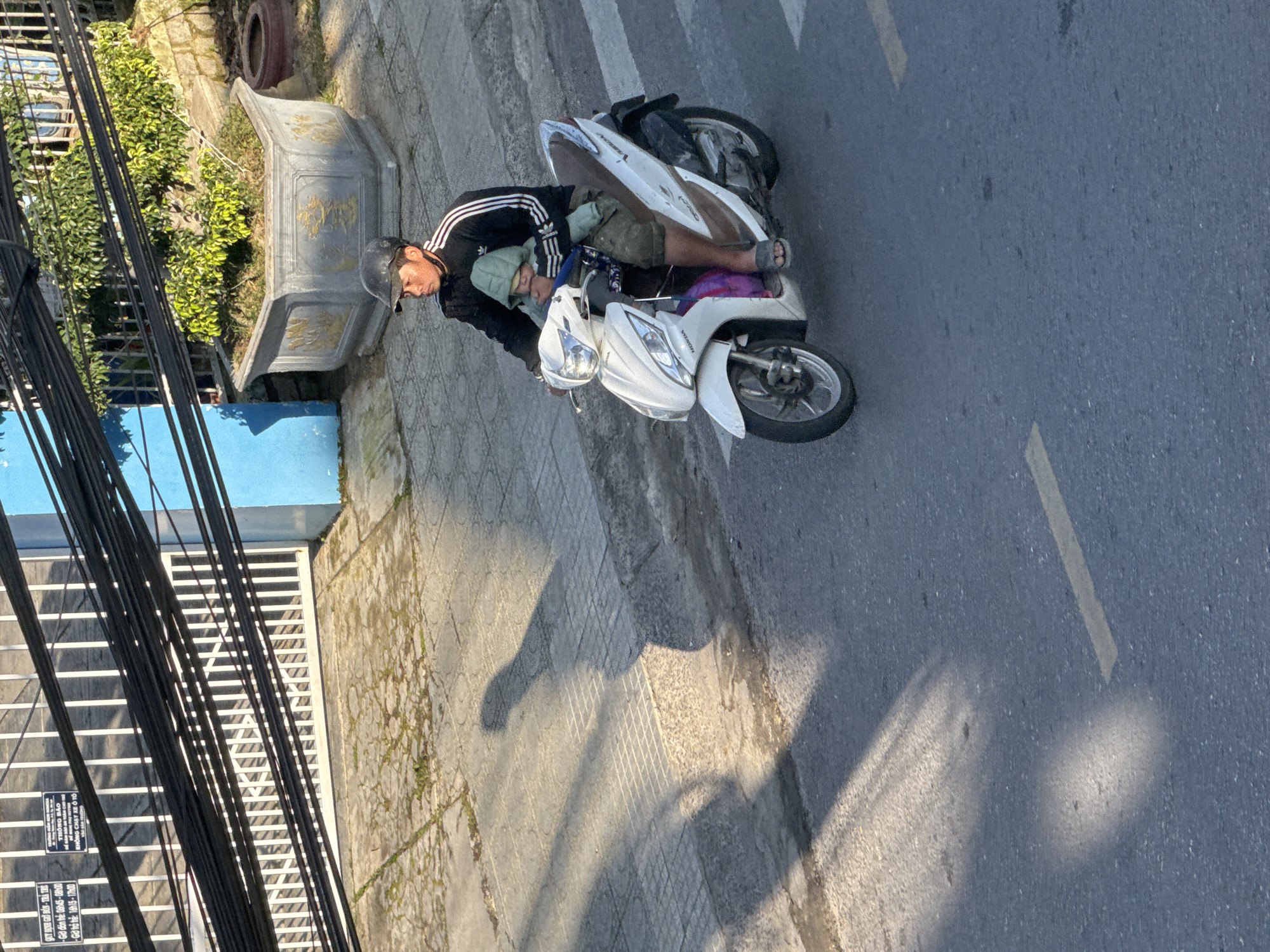 The image captures a street scene in Da Lat, Vietnam, where a man is riding a white Honda scooter with a child seated in front of him. The man wears a dark jacket with white stripes on the sleeves and a dark baseball cap. The child is dressed in a hooded green jacket. They are positioned on a paved road, which appears to be a street in Da Lat. The background reveals a sidewalk with tiled paving, a blue building, a decorative concrete planter with a plant, and a white metal fence with vertical bars. Overhead, a tangle of black electrical wires is visible, casting shadows on the scene. The overall impression is a snapshot of everyday life in the city, highlighting transportation and urban elements.