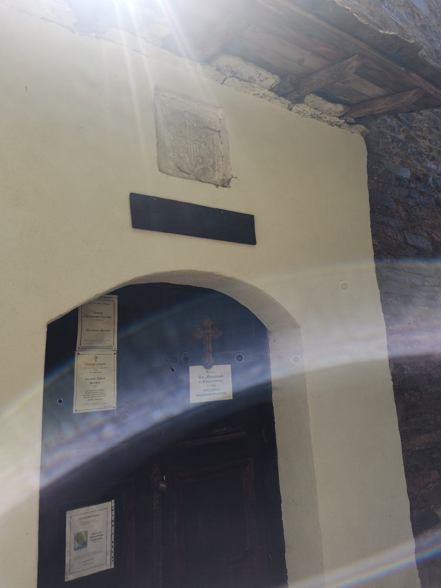 The image captures the entrance of the "Hram Sv. Nikolay" (St. Nicholas Church) in Kovachevitsa, Bulgaria. The building has a light-colored, possibly whitewashed facade next to a stone wall. A stone carving is set above the doorway, with a dark rectangular plaque below it. Several notices are posted near the door, with Bulgarian text. The church entrance is arched, with a metal cross above the door. A sign on the door reads "Храм Св. Николай с. Ковачевица с под постоянно видеонаблюдение," indicating the church's name and that it has continuous video surveillance. There are several areas on the wall that appear to be bullet holes. The upper portion of the facade has visible wood supports. The image is strongly affected by lens flare from the sun.