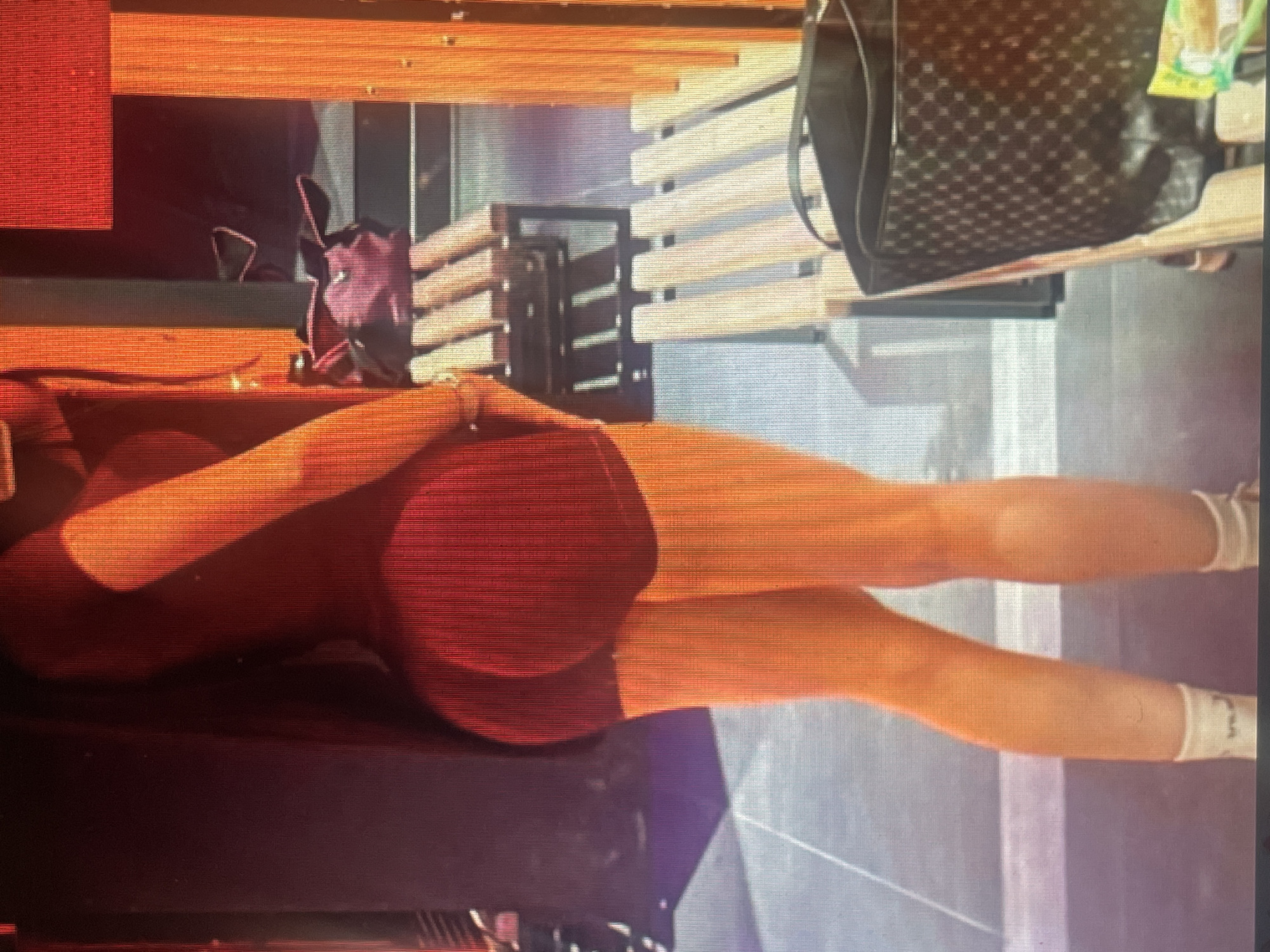 The image shows the lower body of a person wearing a dark red, form-fitting outfit, possibly athletic wear, consisting of a leotard and shorts. The person is facing away from the camera, with their back and legs visible. They are wearing white socks. The background appears to be an indoor or outdoor recreational area with wooden benches and orange structural elements. A dark patterned bag with a handle is visible on one of the benches. The lighting suggests it might be daytime, with shadows cast by the benches. There is no indication of the city of Mantov, Czechia.