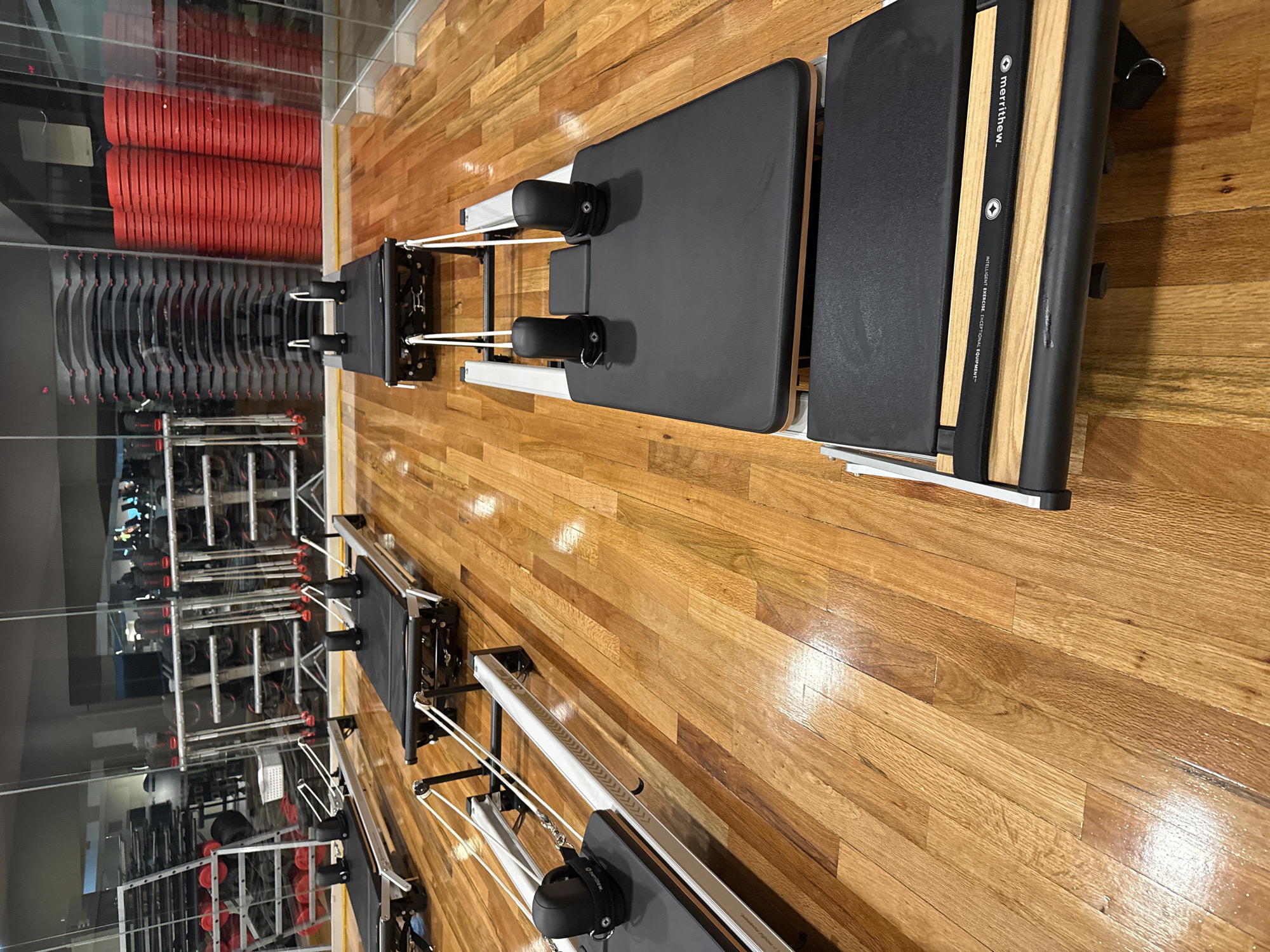 The image shows the interior of a fitness studio. Two Pilates reformer machines are positioned on a hardwood floor with a light brown finish. One machine is closer to the camera and angled diagonally, while the other is further away and more aligned with the vertical axis. A third machine is partially visible in the lower-left corner.
A large mirror covers the left side of the scene, reflecting additional exercise equipment, including stacked Pilates mats and racks holding weights or other equipment. The reflections create a sense of depth and suggest the studio is well-equipped.
Based on the visual information, it is impossible to confirm the location of this image is Puebla, Mexico. The studio appears to be clean and well-maintained. There are no people visible in the frame.