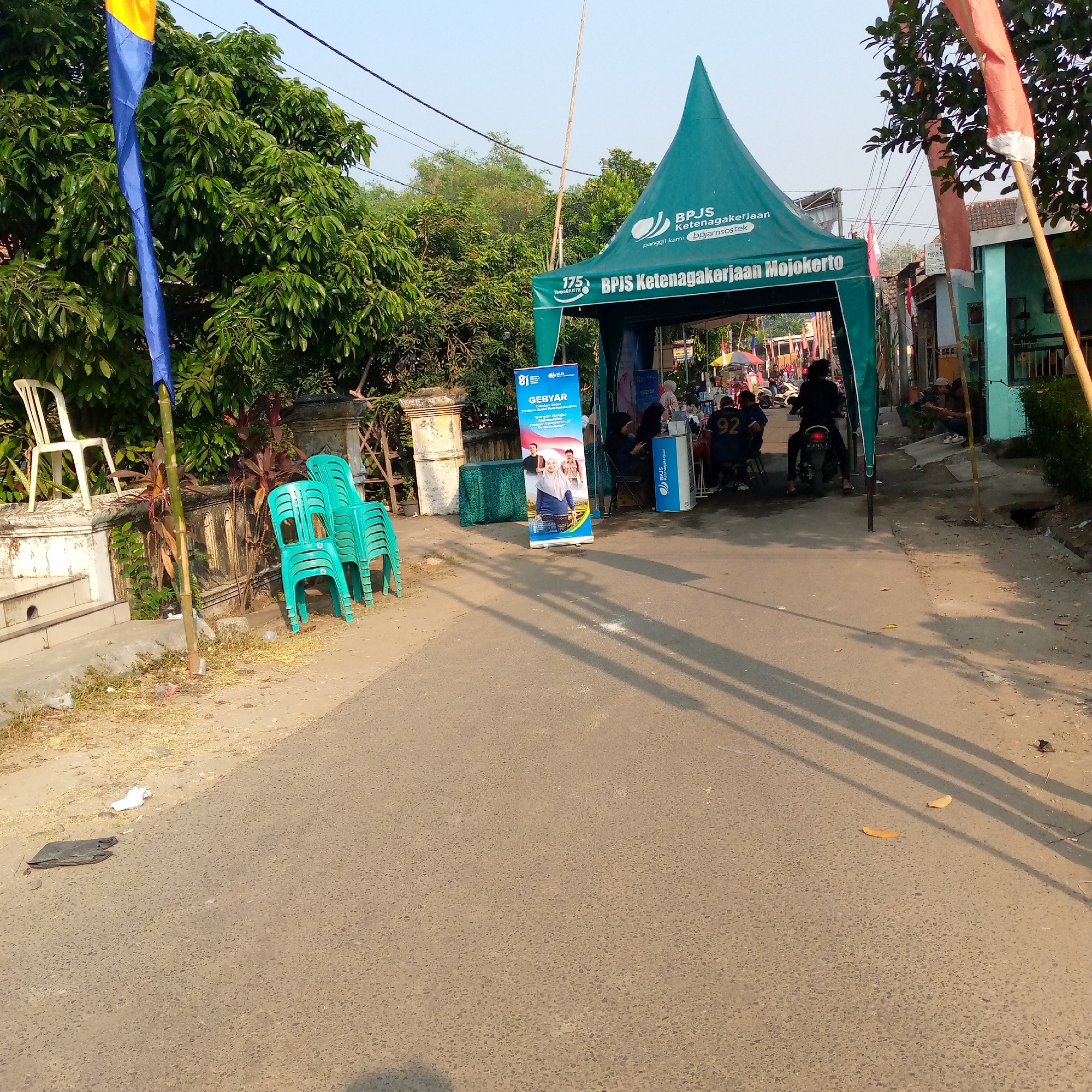 In Tambakagung, Indonesia, an outdoor event is taking place under a green tent with the logo "BPJS Ketenagakerjaan Mojokerto". The tent serves as a booth, with a banner displayed that says "GEBYAR" and features images of people. Several stacks of turquoise plastic chairs are placed on the side of the road, with one white plastic chair also visible on a raised platform. A blue and yellow flag is planted near the chairs. In the background, trees line the street, and buildings with red and blue accents are visible. People are gathered around the tent, with some seated and others standing. A person is riding a motorcycle in the middle of the road. Shadows of people and objects are cast on the asphalt road, indicating a sunny day. The scene appears to be a street in a residential or commercial area, possibly a local gathering or public service event.