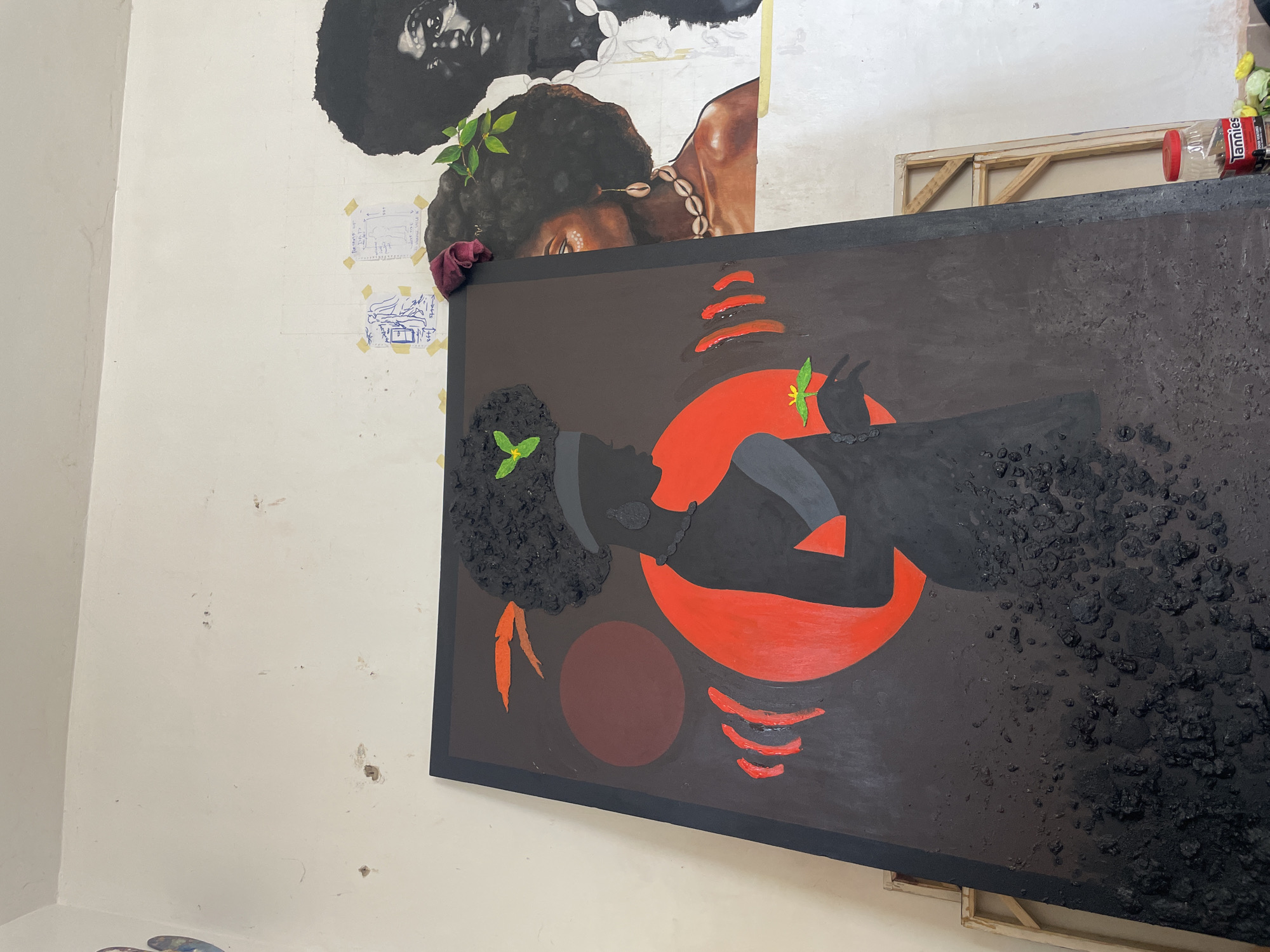 The image shows an indoor art studio in Ado, Nigeria. A large, framed canvas depicting a silhouetted figure of a Black person against a red circular background is prominently displayed. The figure has textured black hair with a green leaf accent. The painting incorporates red, brown, and black hues, and features a dark background with textured elements. Above and behind this painting, partial views of other art pieces are visible, including portraits of Black individuals. The wall has sketches taped to it. A can of "Lannies" sits on top of the framed painting.