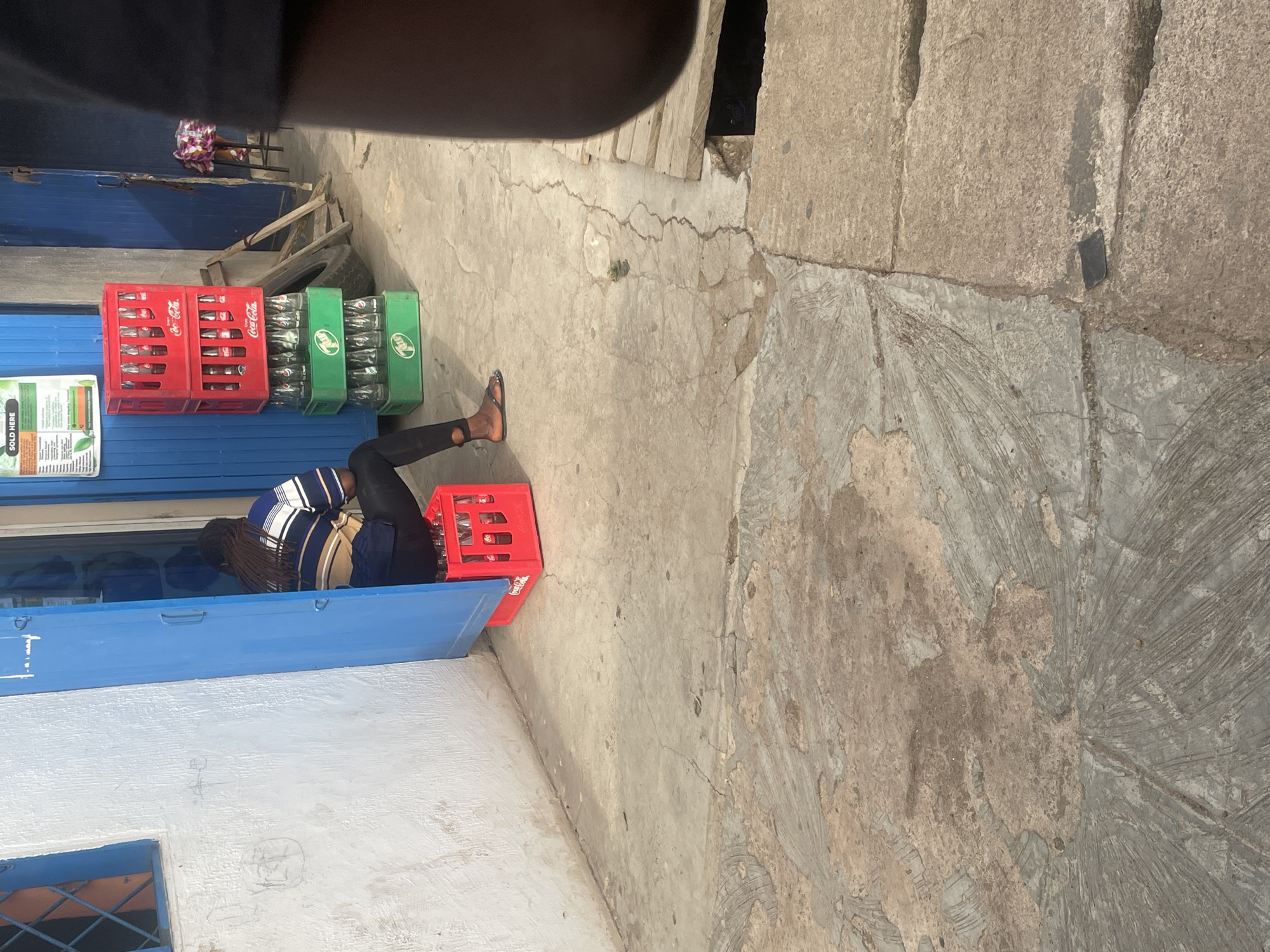 The image, taken in Jos, Nigeria, depicts an outdoor scene focusing on a person engaging with a small street stall or counter. The primary subject, with braided hair and wearing a striped blue, white, and tan top with dark pants, is bent over a bright blue counter, reaching inside or arranging items. To the right of this person, two red plastic crates, clearly labeled "Coca-Cola," are stacked, filled with glass bottles. Next to them are three green plastic crates, also stacked and holding glass bottles, likely another soft drink brand like 7UP. A white sign with green text, partially visible on the blue counter, reads "SOLD HERE."
The ground is a textured, cracked concrete surface, showing signs of wear. Further into the background, another person is partially visible, sitting on a low stool or chair next to a blue wall, wearing a patterned dress. Nearby, a discarded tire and some wooden planks are visible. The surrounding structures consist of buildings with blue and white painted walls, including a small window covered by security bars. The scene is brightly lit, indicating it is daytime. A dark, out-of-focus element from the immediate foreground (possibly the photographer's arm or shoulder) partially obscures the top left portion of the image. The overall impression is one of a busy, informal commercial setup common in local streets.