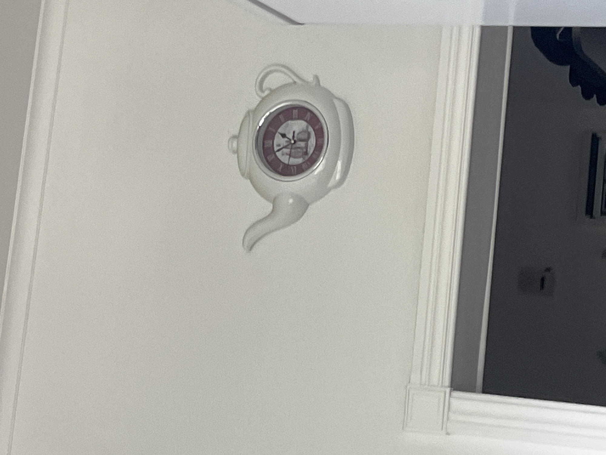 The image is a close-up shot of a wall-mounted clock shaped like a white teapot. The clock has a dark purple face with Roman numerals and decorative hands. The teapot's handle is at the top, and the spout is pointing downwards to the left. The clock is positioned on a plain white wall, with decorative molding visible on the right side of the frame, suggesting an indoor setting. The lighting appears to be artificial and somewhat dim, casting subtle shadows. The overall scene is still and domestic. No people are visible in the image.