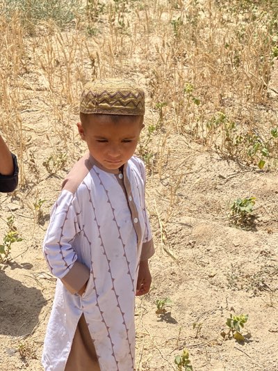 A young boy stands in a dry, sparsely vegetated area in Mir Ahmad Kalay, Afghanistan. He wears a traditional Muslim prayer cap and a white tunic with brown vertical lines and cuffs, with one hand in his pocket. The background consists of dry grass, bare earth, and a few small green plants. A small portion of another person's arm can be seen on the left side of the image. The overall environment suggests a rural setting under bright sunlight.