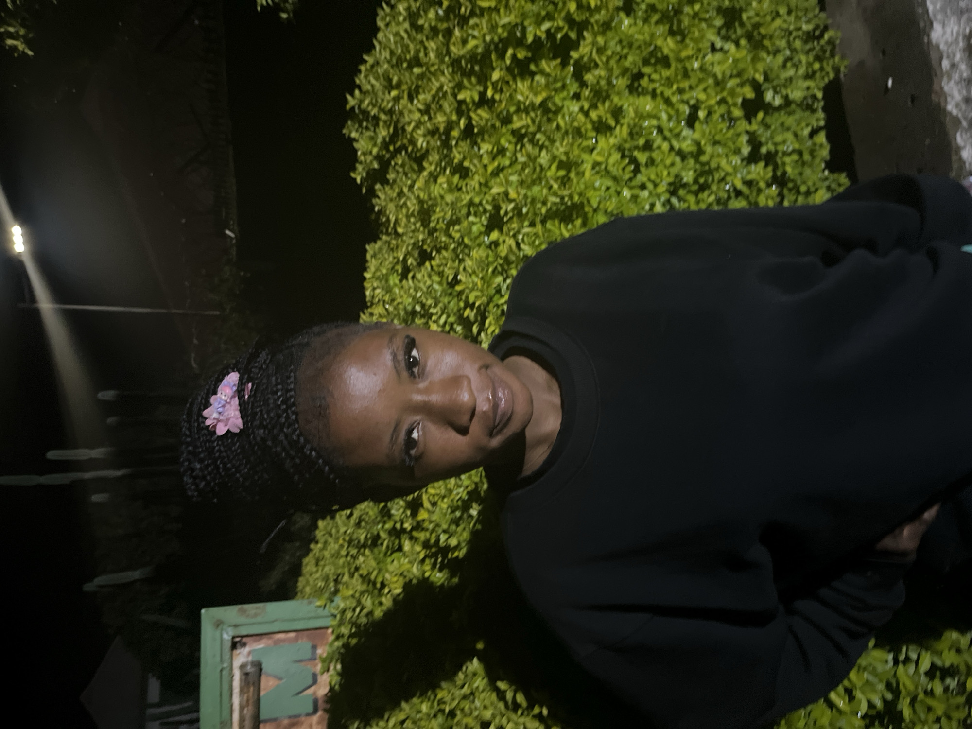 A close-up, waist-up, outdoor portrait of a young Black woman at night. She has braided hair pulled into a high bun, adorned with a pink floral clip. She is wearing a black crewneck sweatshirt and has on makeup, including visible eyeliner and lip gloss. Her left hand is resting on her hip, with a hint of teal fabric visible at the bottom of the frame.
Behind her, a dense, well-maintained green hedge provides a backdrop, illuminated by a bright light source to the upper right. To the left, a green sign with a weathered, possibly metallic letter "M" is partially visible. The ground appears to be asphalt or dark pavement, with a few small white specks that could be debris or reflections. The overall lighting suggests artificial illumination in an urban or semi-urban environment, consistent with nighttime in Jos, Nigeria.