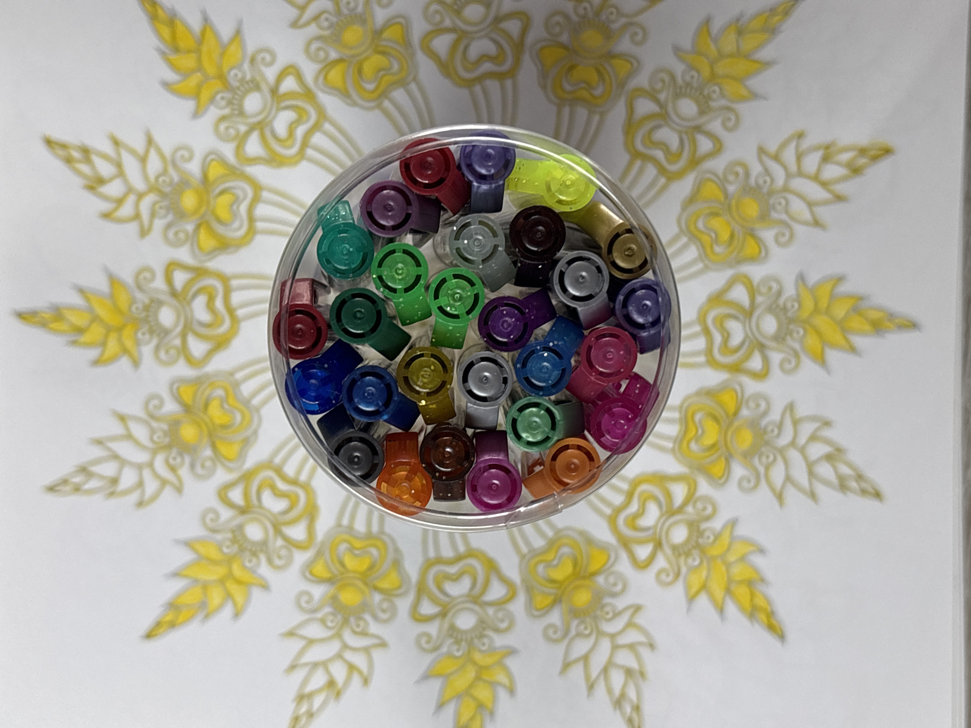 The image shows a clear plastic container filled with colorful pen caps, viewed from directly above. The pens are arranged in a dense cluster within the container. The container is placed on a white surface decorated with a yellow and gold floral pattern, which appears to be a drawing or print. The pattern is symmetrical and radiates outwards from the center. The lighting is bright and even, creating minimal shadows. There are no people or animals present in the image. The background appears to be a flat surface. There is no information in the image to suggest a location in Sochi, Russia.