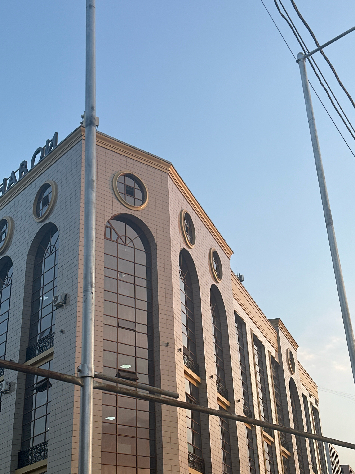The image shows the corner of a building with a light-colored tiled facade. The building has a prominent cornice and features arched windows and round windows. There are metal poles and overhead wires in the foreground and extending into the sky, which is a clear blue. The lighting suggests it is daytime, possibly late afternoon, given the warm tones reflecting on the building's surface. Some of the windows are dark, while others show reflections or appear to have internal lighting. The text "HAPOH" is visible in large blue letters on the top facade of the building.