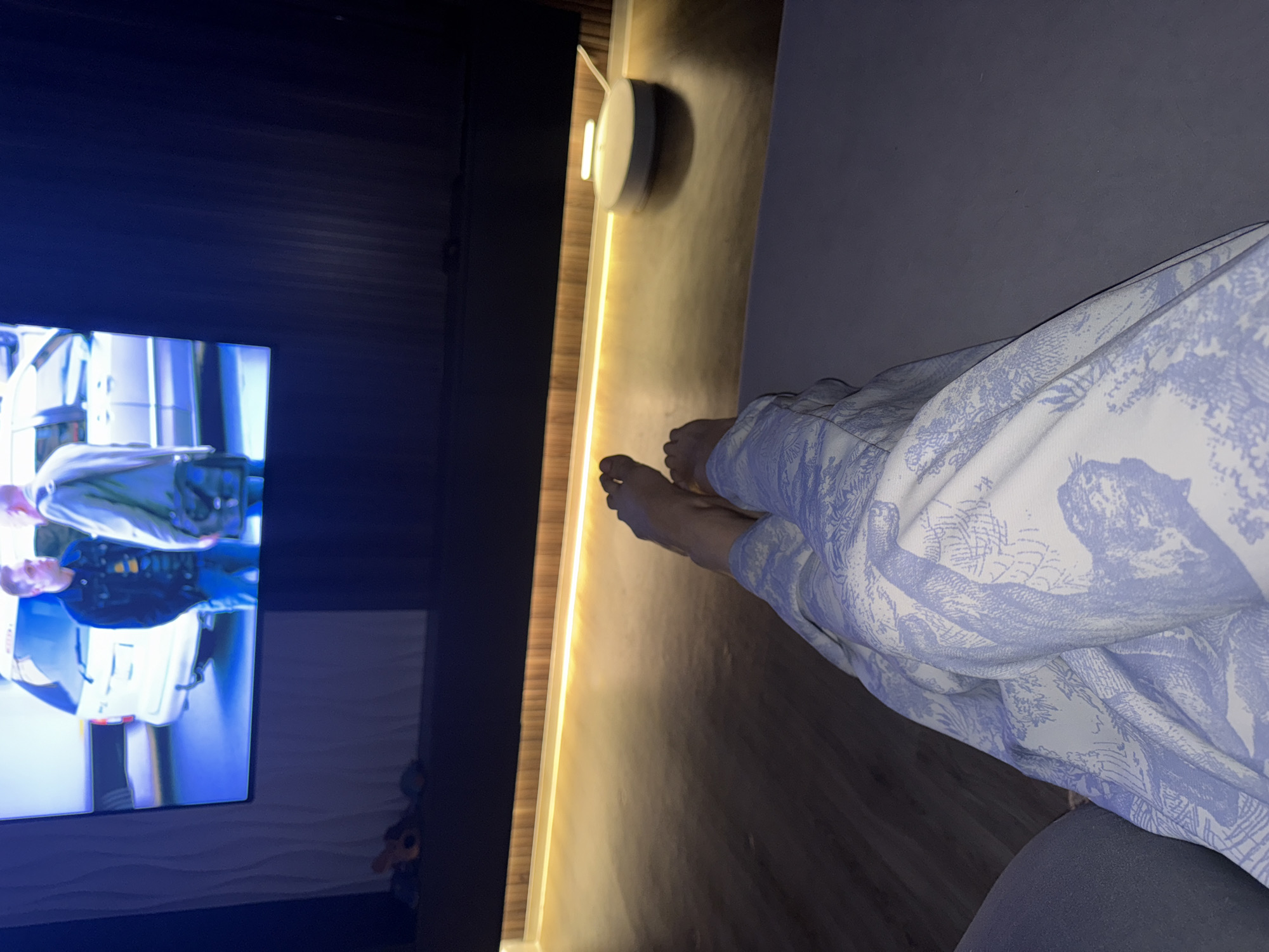 A person is lying on a bed in front of a television in a room. The television is on and shows a scene of people in what appears to be a police setting. The person's feet are visible at the edge of the bed, which is covered in a white and blue patterned blanket. The room is lit by a light fixture behind the bed. There is no indication of the location being Лобаново, Russia.