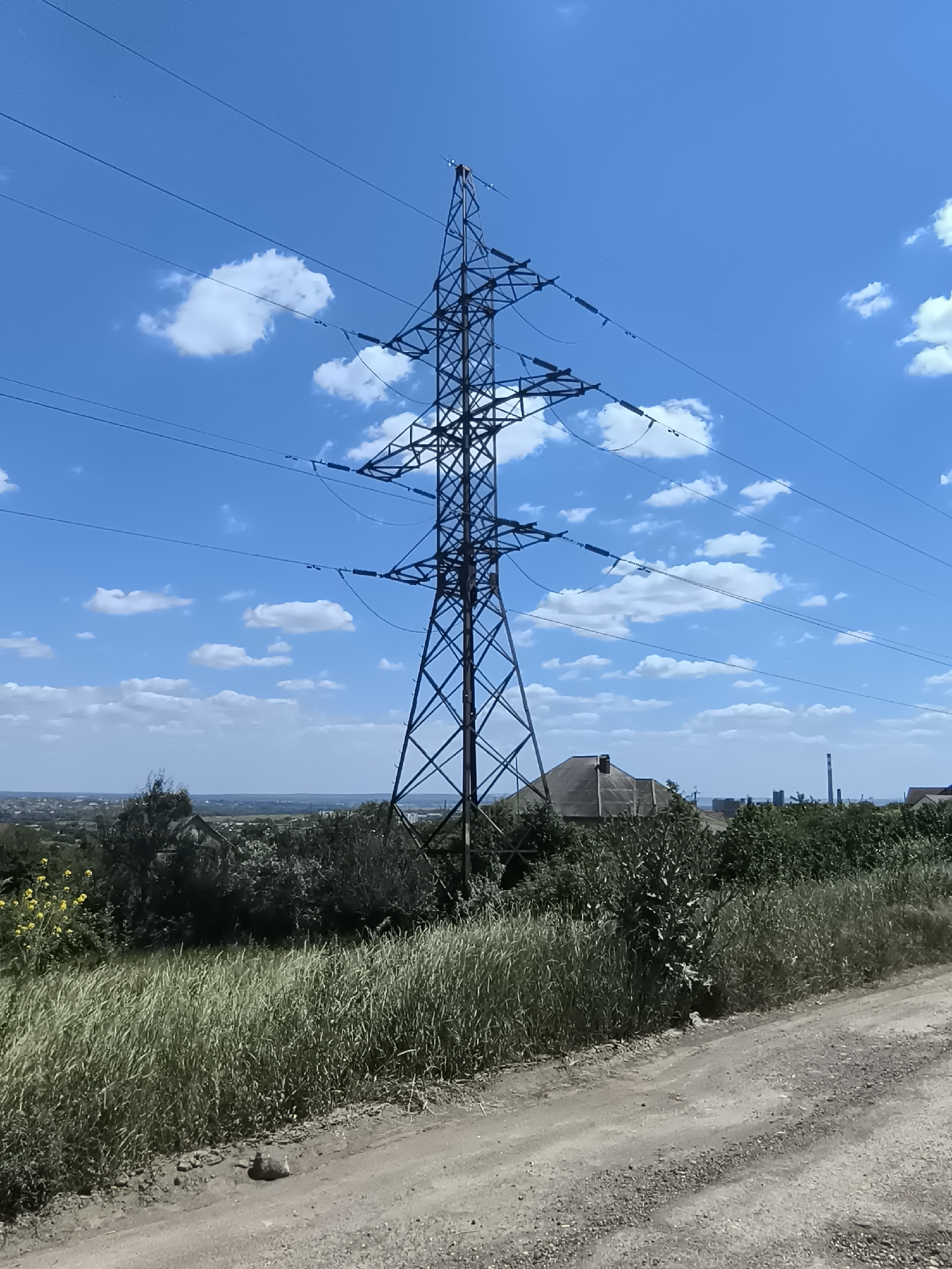 A tall, lattice steel power pylon stands centrally in a landscape under a bright blue sky with scattered white clouds. The pylon is surrounded by trees and vegetation, with a dirt road in the foreground. In the distance, a town or city is visible, with buildings and what appears to be a smokestack or industrial structure. The scene appears to be located in or near Varnița, Moldova.