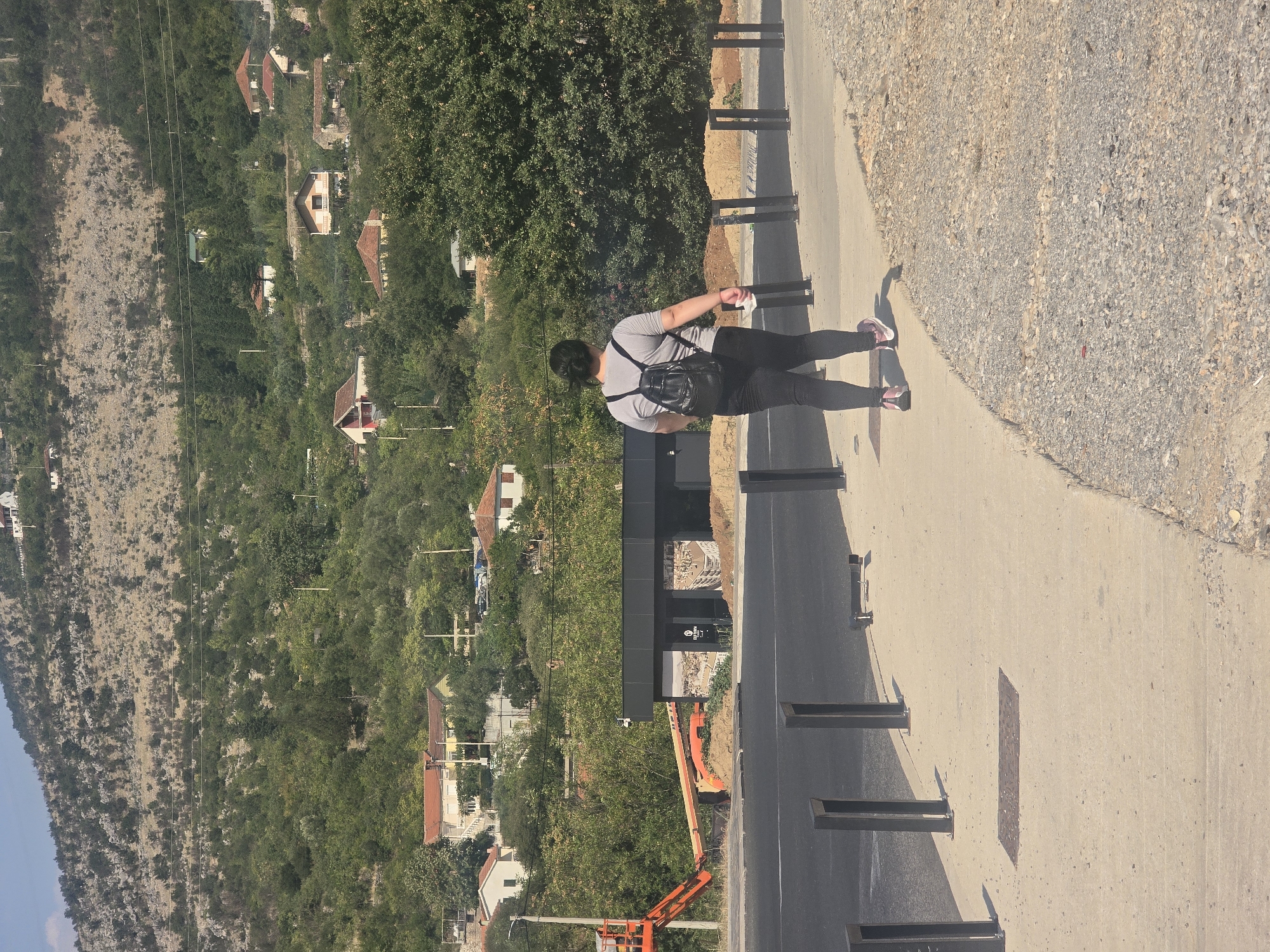 An outdoor, eye-level shot shows a woman from behind walking on a sidewalk next to a road. She has dark hair pulled back into a ponytail, wears a grey t-shirt and black leggings, and carries a black backpack. Her feet are clad in pink and white athletic shoes. The road is dark asphalt. The sidewalk is light concrete, bordered by a gravelly area. Several dark metal posts are spaced along the sidewalk. In the background, a row of dark metal bollards marks the edge of the road. A small, dark-colored building with large windows is visible behind the bollards. To the left of the building, an orange construction lift is parked. Beyond the building and construction area, the landscape rises steeply with a dense covering of green trees and scattered residential buildings with red-tiled roofs. The hillside is rocky and sparsely vegetated higher up. The scene is bathed in sunlight, casting shadows. The location is an area of Podgorica, Montenegro, likely on the outskirts or a hillside district, given the mix of urban infrastructure and the prominent natural landscape.