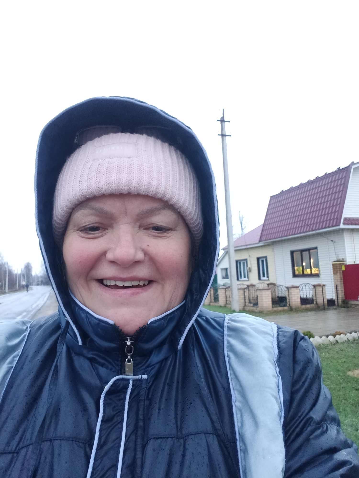 A close-up outdoor selfie depicts a smiling, middle-aged woman wearing a dark blue hooded jacket with light blue/grey accents and a light pink knitted beanie hat. Her expression conveys happiness, with her mouth open in a broad smile, showing her teeth. Small water droplets are visible on her jacket, indicating damp or rainy weather.
The background, set in Красный Маяк, Russia, reveals an overcast, white sky. To the left of the woman, a wet road extends into the distance, flanked by blurred trees and a faint, distant figure. To her right, a house with a distinctive maroon tiled roof and white siding is visible, partially obscured by a low brick fence. A utility pole stands in the mid-ground. Green grass is present in the lower right of the frame. Faint, illegible text appears on a label inside her beanie hat. The overall scene suggests a cool, overcast day in a residential or rural area.