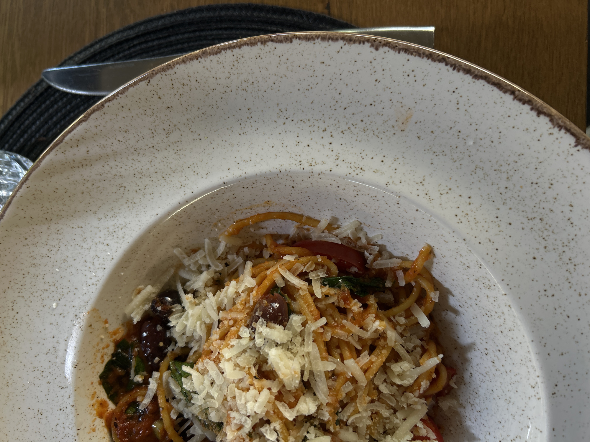 Overhead close-up shot of a partially filled white plate with speckled brown edging. The plate contains spaghetti with tomato sauce, spinach, olives, and shaved parmesan cheese. A silver knife is visible above the plate. The plate sits on a black woven placemat on a wooden surface. The location is possibly Sofia, Bulgaria, but no specific identifying features confirm this. No people are visible. The focus is entirely on the food.