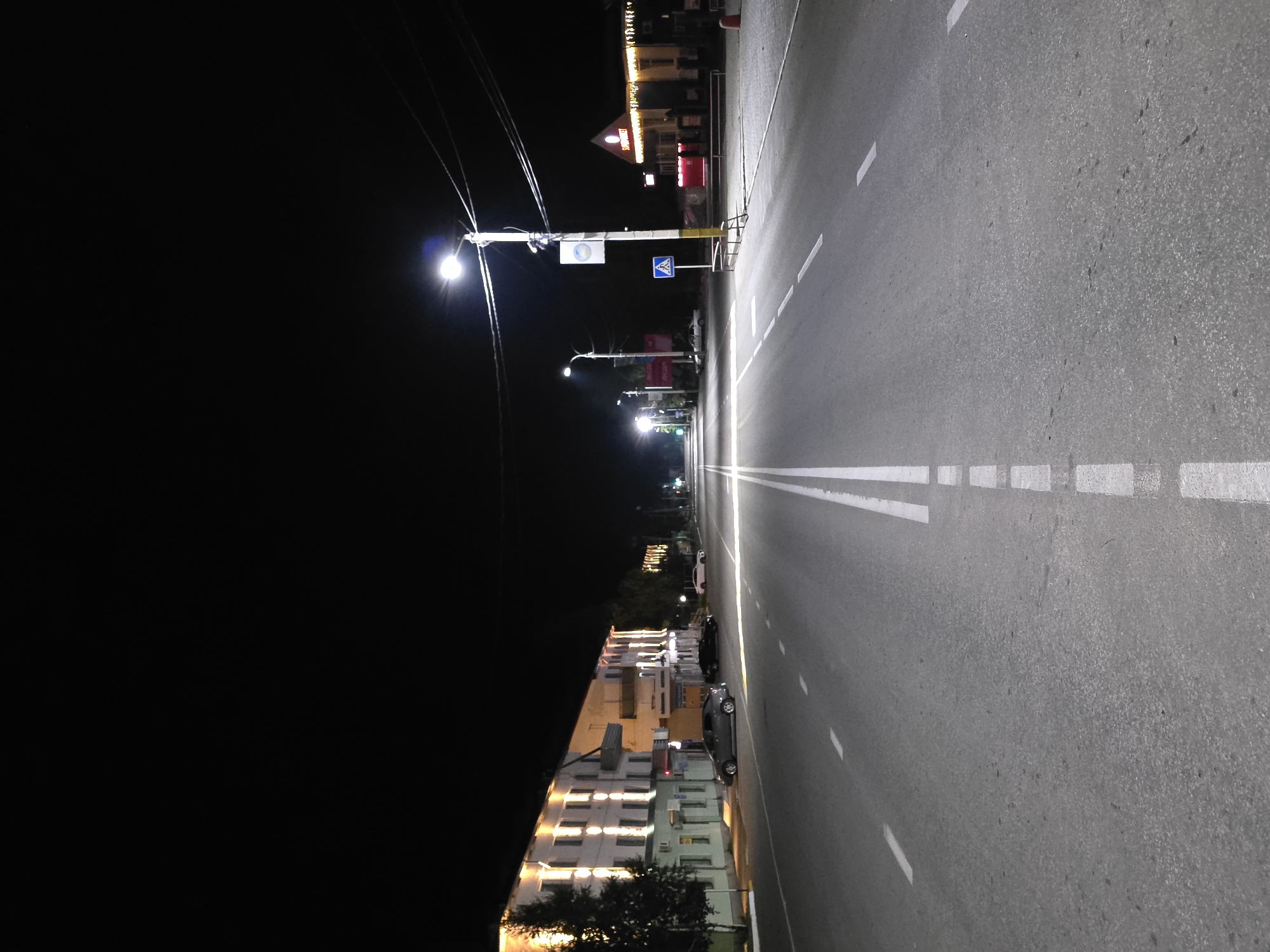 The image shows a street scene at night in Qarabalıq, Kazakhstan. A paved road with white lane markings runs through the center of the frame, extending into the distance. Streetlights illuminate the road, casting a glow on the asphalt. On the left side of the road, a multi-story building with illuminated windows and decorative lights runs along the street. Several cars are parked along the side of the road in front of the building. On the right side of the road, there is a building with a sign that reads "SUPERMARKET". A traffic sign indicating a pedestrian crossing is visible near the supermarket. Power lines and street lamps are also present throughout the scene. The overall environment is dark, typical of a street at night.