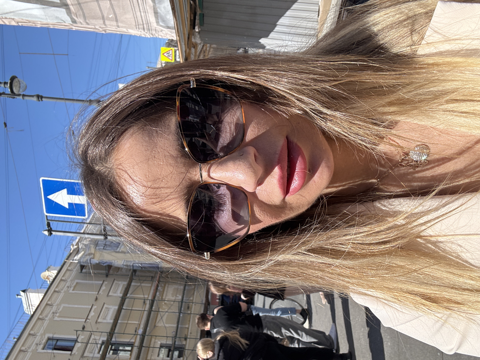 The image is a close-up, selfie-style portrait of a woman with long, blonde hair, wearing large, tortoiseshell sunglasses and a light-colored top. Her lips are painted a vibrant pink. A delicate necklace with a sparkling pendant is visible on her décolletage. The background is slightly out of focus, but it depicts an urban street scene.
In the background, there are buildings with light-colored facades and visible scaffolding. A blue traffic sign with a white arrow pointing to the left is mounted on a pole. Another yellow triangular traffic sign with a warning symbol is also visible. Several people are walking on the street, some of whom are partially visible. The sky is clear and blue, suggesting a sunny day. The sunlight casts shadows, indicating it is likely daytime. The scene appears to be set in Saint Petersburg, Russia, given the architectural style and traffic signs. The woman's pose and direct gaze into the camera suggest she is taking a selfie. There are no discernible emotions conveyed beyond a sense of self-presentation. The sunglasses have the brand name "LANEL" subtly inscribed on the lens.