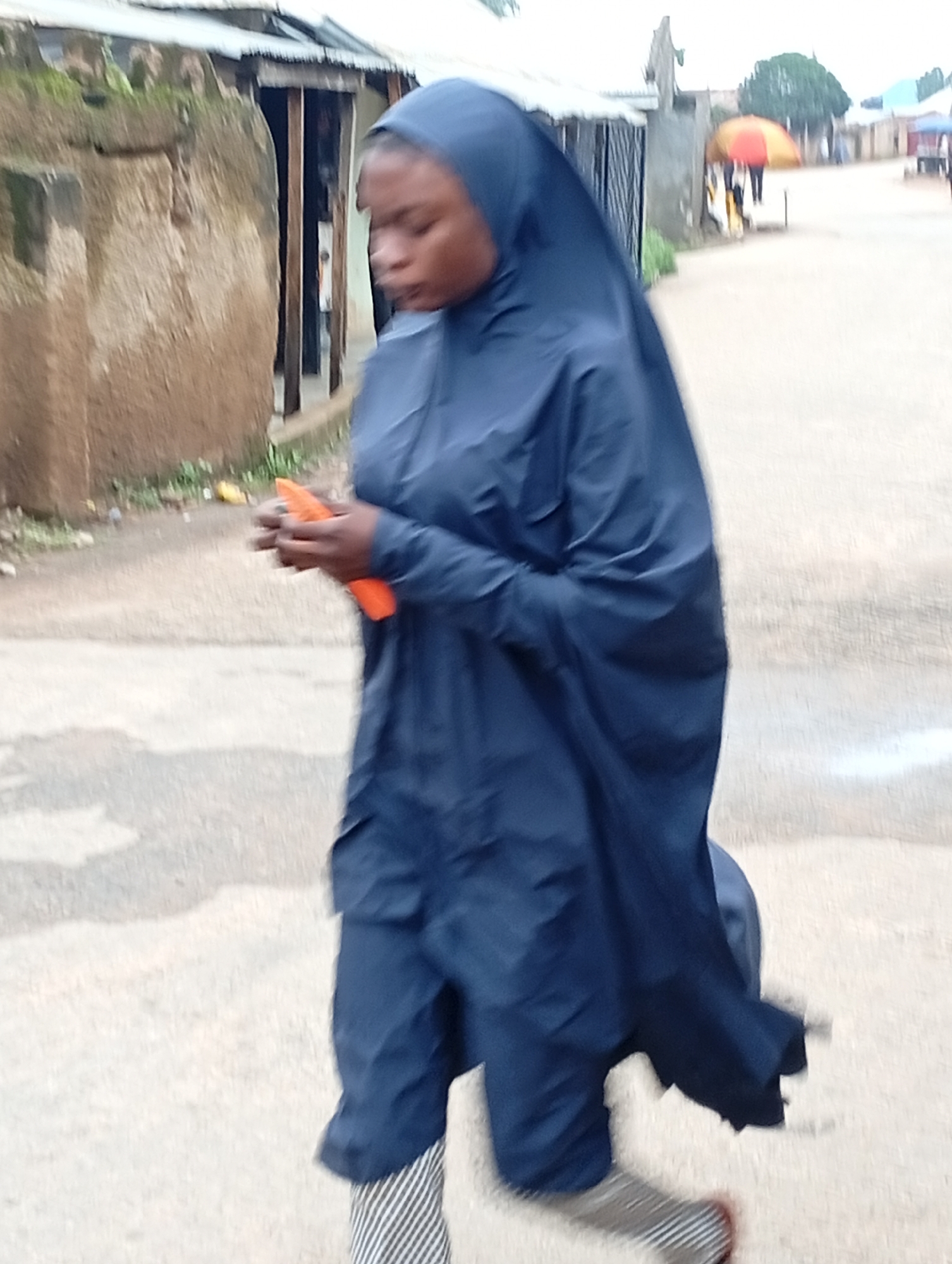 A person wearing a dark blue hijab and a matching full-length garment, along with striped lower leg coverings, walks on a paved street in Jos, Nigeria. The individual is holding a bright orange object, possibly a phone, in their right hand. To the left of the person is a rough, textured wall, suggesting a building's facade. In the background, a street stretches ahead with buildings and a few individuals, one holding a red and yellow umbrella. The sky appears overcast, indicating a potentially cloudy or rainy day.