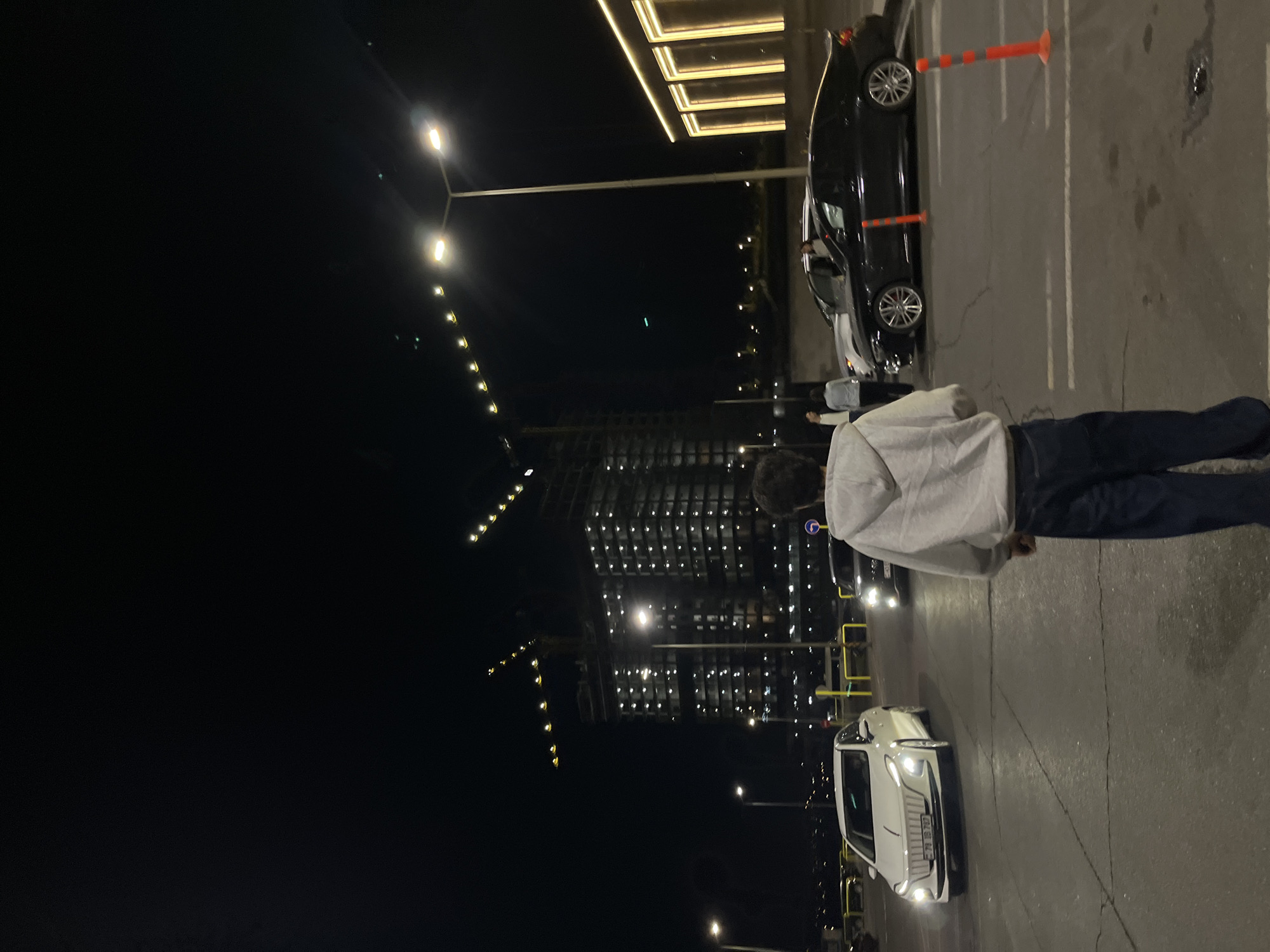 At night in Yerevan, Armenia, a person wearing a gray hoodie and blue jeans stands in a parking lot. A black car is positioned at an angle near orange cones, and a white car faces the viewer, illuminating the foreground. Streetlights and the lighted windows of a large building illuminate the scene.