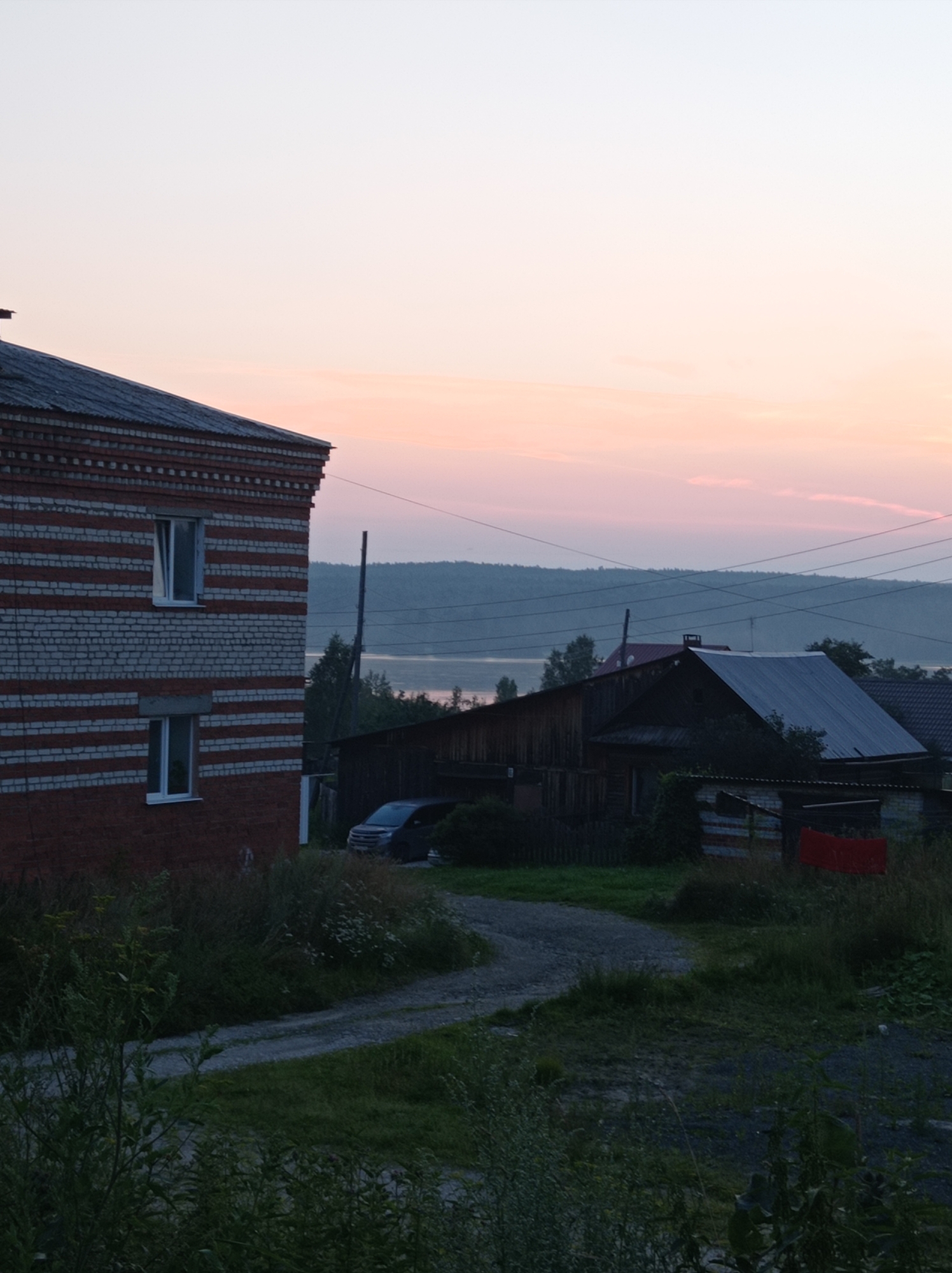 The image depicts a rural landscape at dusk, likely in the city of Degtyarsk, Russia, given the architectural style and vegetation. In the foreground on the left, a two-story brick building with alternating red and white stripes is visible. It has several windows, some of which are open. To the right of this building, a dirt road curves through overgrown grass and weeds. Further back, several wooden structures, possibly houses or sheds, are present, with dark roofs. One of these structures has a light-colored metal roof. A dark-colored car is parked on the dirt road near the wooden buildings. In the background, a body of water is visible, bordered by a tree-covered hill or forest. Power lines are strung across the middle ground, and the sky above is a gradient of pale orange and pink, indicating sunset or sunrise. There are no people visible in the scene. The overall impression is one of a quiet, somewhat rustic, rural setting during twilight.