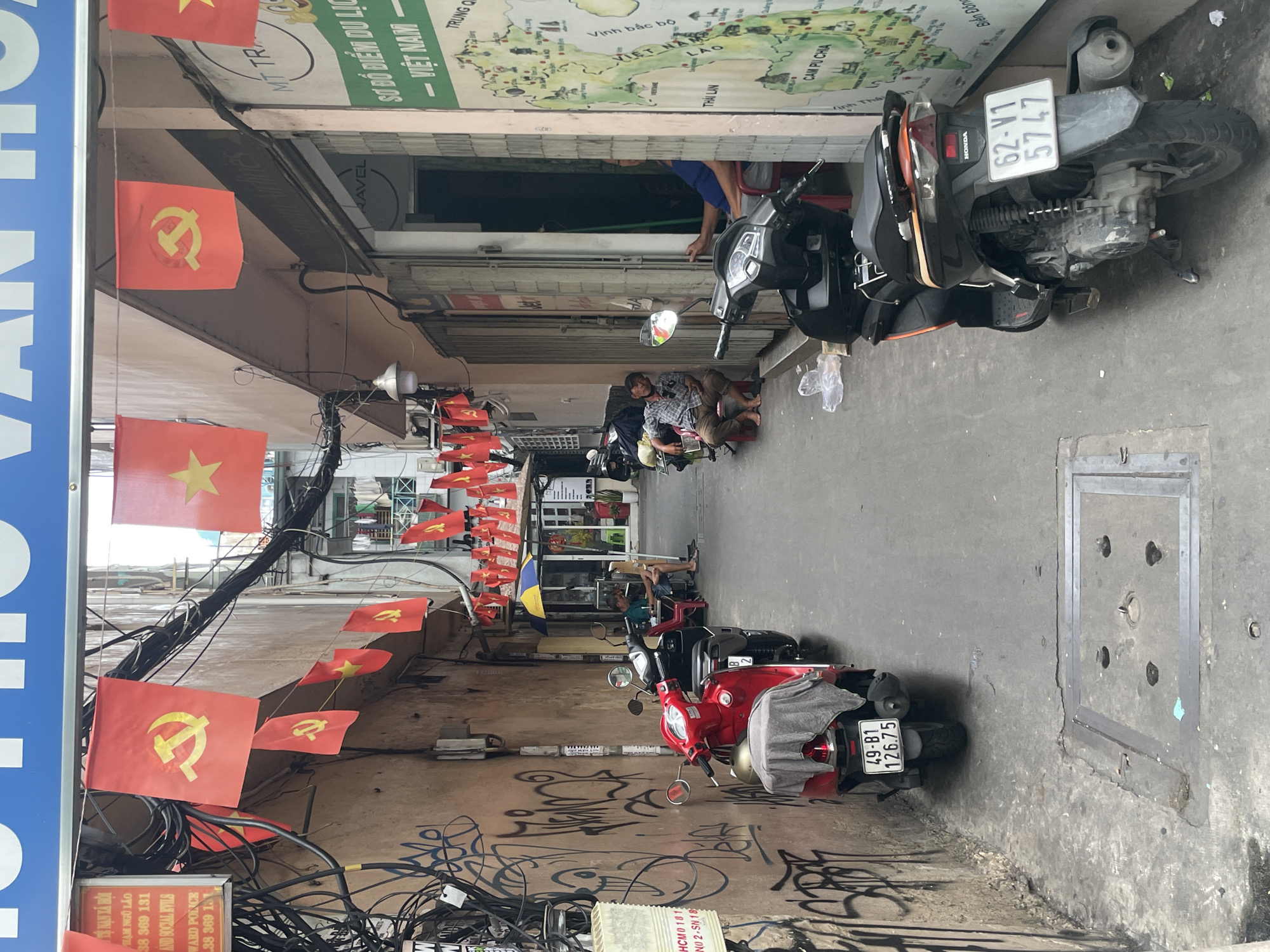 The image depicts a narrow alleyway in Thủ Đức City, Vietnam. The alley is lined with buildings and features numerous red flags with yellow stars, a symbol of Vietnam, and also red flags with a yellow hammer and sickle, symbols of communism, strung overhead. Several motorbikes are parked along the sides of the alley. In the foreground, a red motorbike is parked, and further down, a black motorbike is visible. People are seated on red stools, some appearing to rest or converse. Graffiti covers the wall on the left side of the alley. A large map of Vietnam is displayed on the wall of a building on the right. The environment appears to be urban, with power lines and electrical equipment visible. The overall scene suggests a bustling, everyday scene in a Vietnamese city.