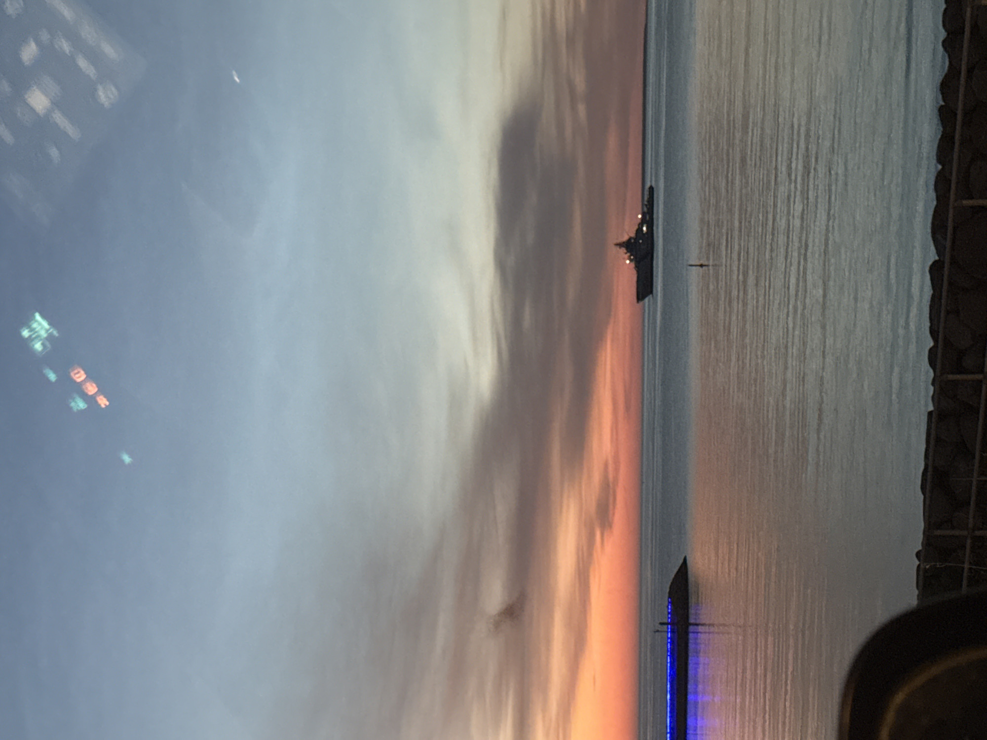 The image captures a serene view of the Baltic Sea at dusk, likely from the coast of Tallinn, Estonia. The sky displays a gradient of colors, transitioning from a soft orange and pink near the horizon to a pale blue overhead, indicative of a sunset or sunrise. A prominent element in the scene is a large naval ship, silhouetted against the horizon and illuminated with lights. In the foreground to the left, a pier or breakwater is visible, adorned with bright blue lights, reflecting on the calm water. The water itself is placid, with gentle ripples. The image also shows a hint of greenery or a fence-like structure at the bottom edge, suggesting a waterfront promenade or park. Above, reflections of lights or possibly an electronic display are visible on a surface, creating abstract shapes in the sky. There are no people clearly depicted in the image.