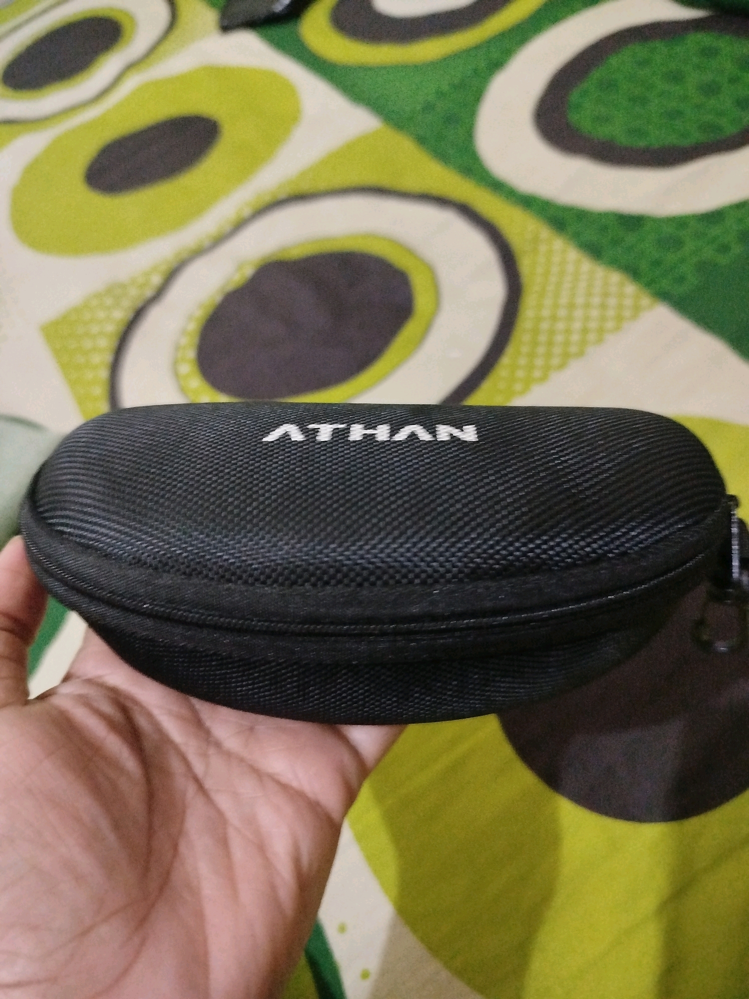 A black oval glasses case with "ATHAN" written in white on top rests on a patterned surface. The surface is a fabric with abstract oval shapes in green, white, and gray. A hand is holding the glasses case from the bottom left. The image is a close-up shot.