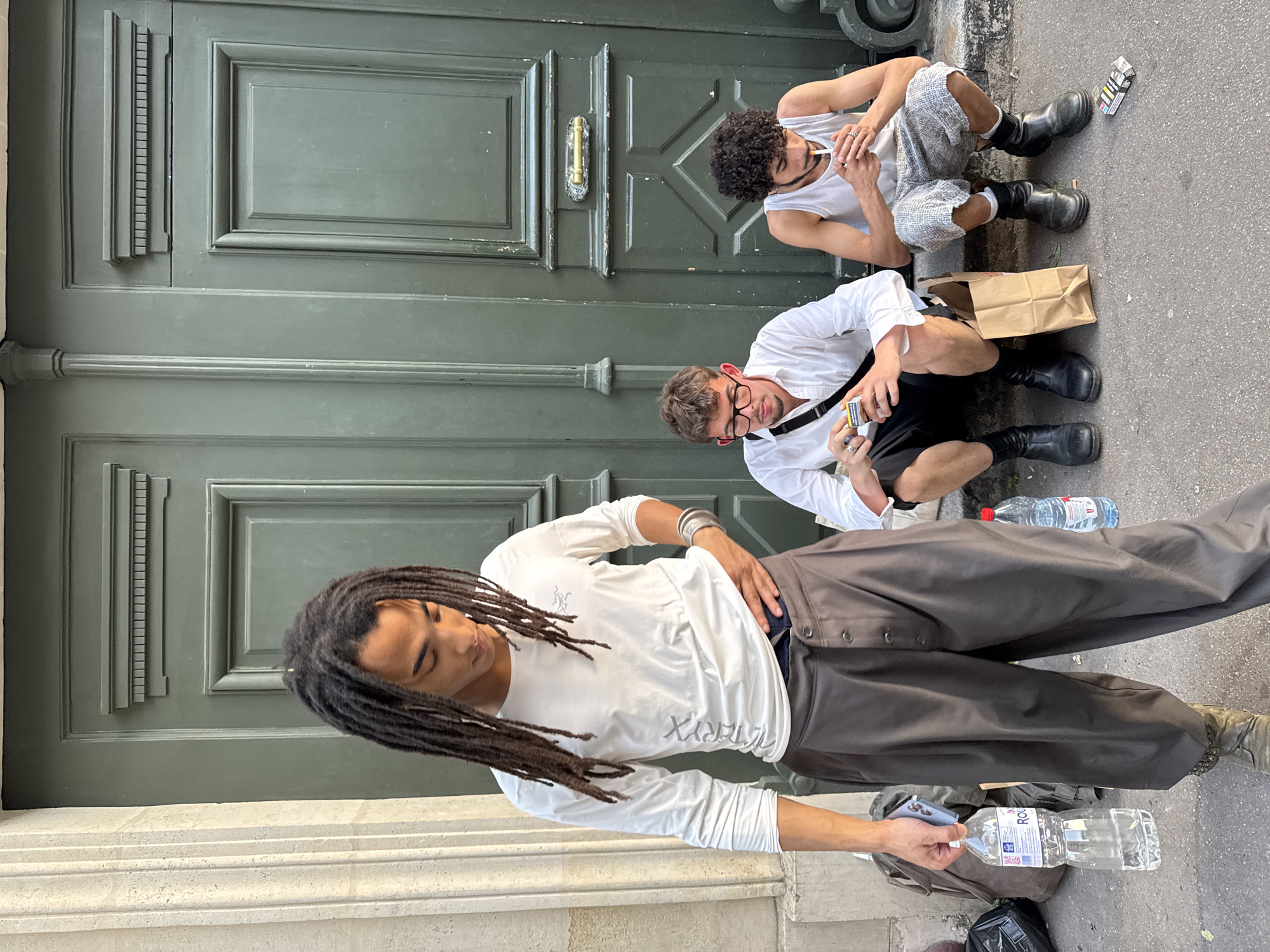 Three young men are seated on the pavement next to a large green door. One man with dreadlocks is standing and holding a water bottle. The door appears to be a traditional Parisian door, suggesting the location is in Paris, France. The standing man wears a white long-sleeved shirt and wide, dark-colored pants. One seated man is wearing a white shirt and black shorts, and the other is in a white tank top and light-colored shorts. Both seated men have dark boots. A paper bag and a pack of cigarettes are on the pavement near the seated men. Water bottles are also present.