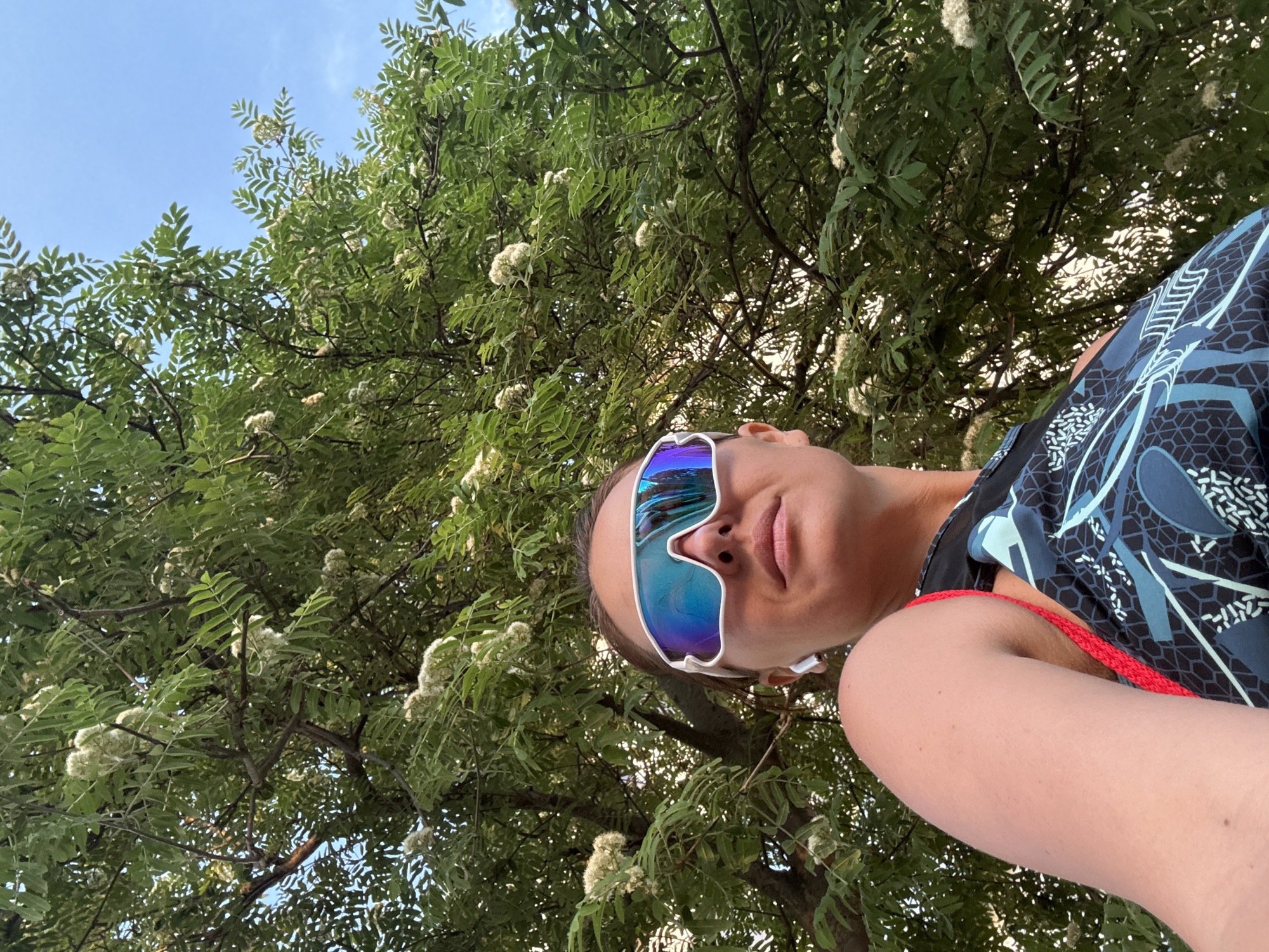 A low-angle medium shot shows a person wearing reflective sunglasses, a blue-patterned shirt, and a red strap over their shoulder. The person is standing beneath a tree with green leaves and white flowers. The background also includes a patch of blue sky. The image was possibly taken in a park or wooded area, but further location context beyond what is shown cannot be determined from the provided picture, especially to place in the city of Chaikovsky, Russia.