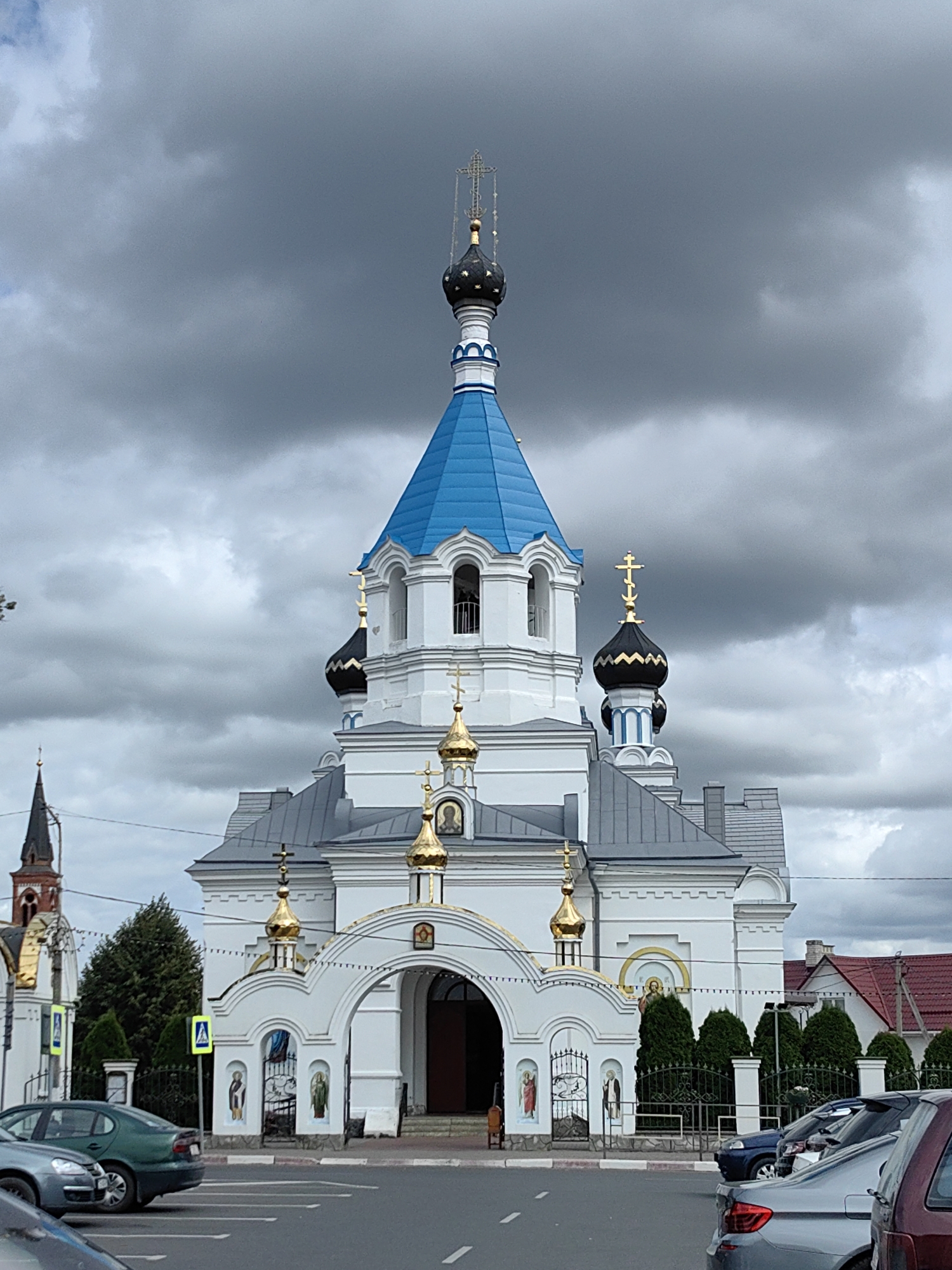 This is a full-frame, eye-level, outdoor photograph taken on an overcast day, showing the Resurrection Church in Pastavy, Belarus. The church is the primary subject, a white, multi-tiered building with a prominent bright blue dome and several smaller, gold-capped domes topped with crosses. The church features white arched entrances and decorative golden accents. To the left of the main church, a portion of another building with a dark spire is visible, also featuring gold elements. In the foreground, a paved road with parking lines is present, with several parked cars visible. These include a dark green sedan, a gray sedan, and the rear of a maroon SUV. A black metal fence with conical green trees behind it runs along the sidewalk in front of the church. Two yellow pedestrian crossing signs are also visible. The sky is heavily clouded with dark gray storm clouds.
