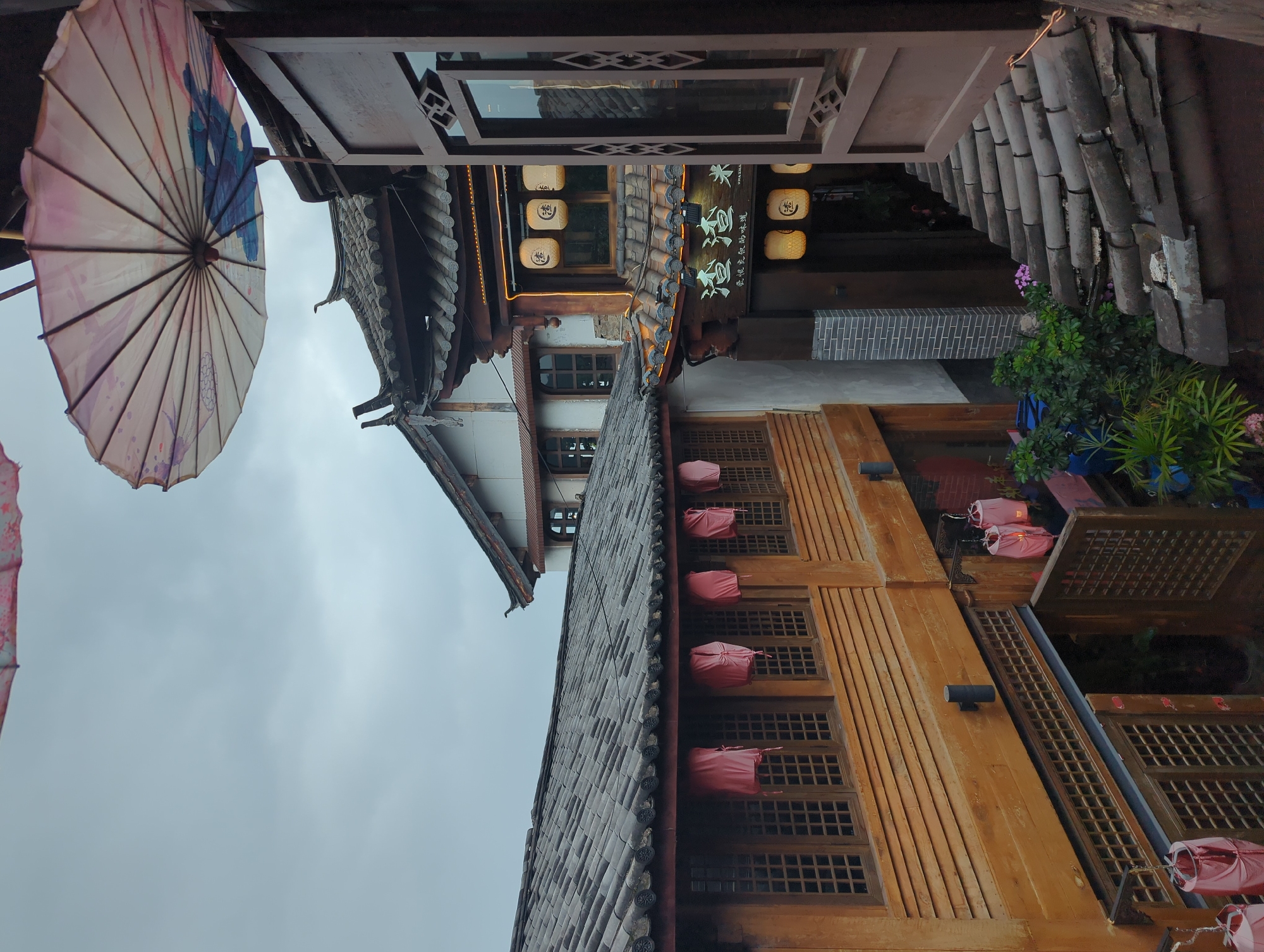 This is a medium-shot, eye-level, outdoor photograph taken in the Gucheng District, China. The image features traditional Chinese architecture with dark tiled roofs and wooden structures. Several pink lanterns are hung along the wooden balconies and over an open doorway. An open wooden window frame occupies the right side of the foreground, through which more of the buildings and a tiled roof are visible. A decorative, patterned umbrella is suspended in the upper right portion of the frame. The sky is overcast and gray. No people are visible in the scene. The overall impression is one of an atmospheric, historic streetscape in a Chinese city.