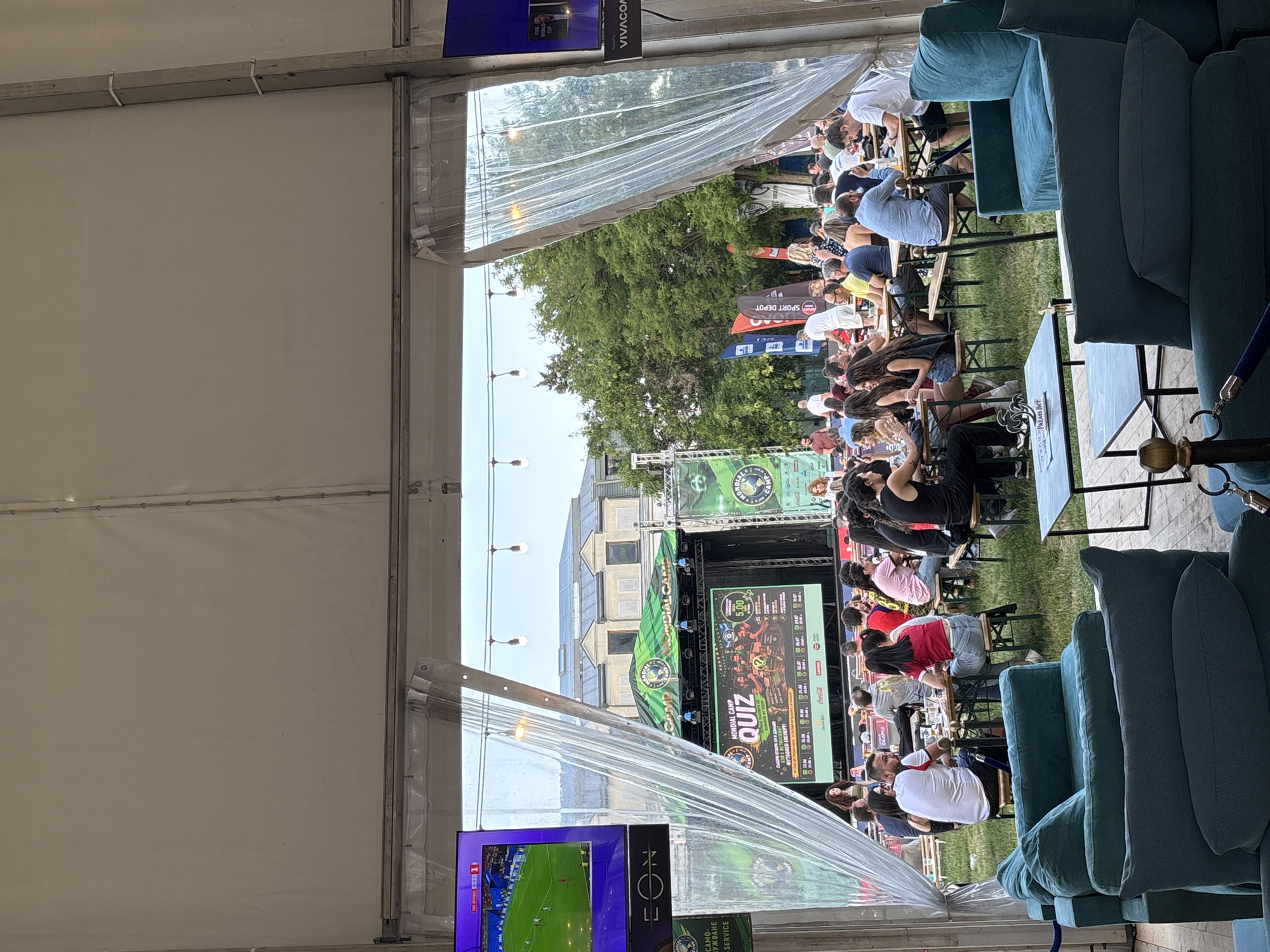 This is a wide shot of an outdoor event in Sofia, Bulgaria, likely taking place during a football match as indicated by the screens displaying a football game and the "Mondial Camp" branding. The event is set up under a large white tent, with clear plastic sheeting acting as partial walls, revealing the outside. In the foreground, there are comfortable teal-colored sofas arranged around low tables.
Further back, beyond the sofas and to the left, a large screen displays a football game with the "EON" logo at the bottom. Center stage, a large screen is set up with a "Mondial Camp QUIZ" advertisement, showing a graphic of people with question marks and what appears to be prize money. Surrounding the stage are various banners and flags, including one with "SPORT DEPOT" and another with "VIVACOM".
In the mid-ground, numerous people are seated at tables and benches on a grassy area, engaged in conversation and watching the event. They are dressed in casual summer clothing. The atmosphere appears relaxed and social. In the background, there are trees and buildings, indicating an urban or semi-urban setting. The lighting suggests it is daytime, possibly late afternoon or early evening.