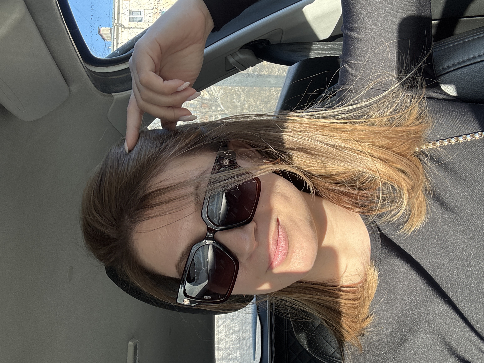 A female individual is depicted in a close-up, seated in the passenger seat of a vehicle in Chaikovsky, Russia. The subject, appearing to be in her 30s-40s, has light brown to blonde hair and wears large, dark, square-framed sunglasses with a gold-colored emblem on the left temple. She is dressed in a dark, possibly black, long-sleeved top with a subtle texture and has a gold-colored chain strap across her left side, indicative of a bag. Her right hand, with light-colored, almond-shaped manicured nails, is raised to touch her hair near her right temple. She looks towards the camera with a subtle smile.
The vehicle's interior features black leather seats with a quilted design, a light gray ceiling, and a visible sun visor above the passenger window. Strong sunlight enters from the passenger side, illuminating the subject and casting shadows.
Through the upper right portion of the passenger side window, a clear blue sky is visible above a light-colored, multi-story building, likely a residential or commercial structure. This building features multiple windows and a prominent dark chimney or vent on its rooftop. Small specks or droplets are present on the exterior of the window glass. A blurred urban background with bare trees is visible through other windows, consistent with an outdoor setting in Chaikovsky, Russia. The reflection in the sunglasses shows the vehicle's interior.