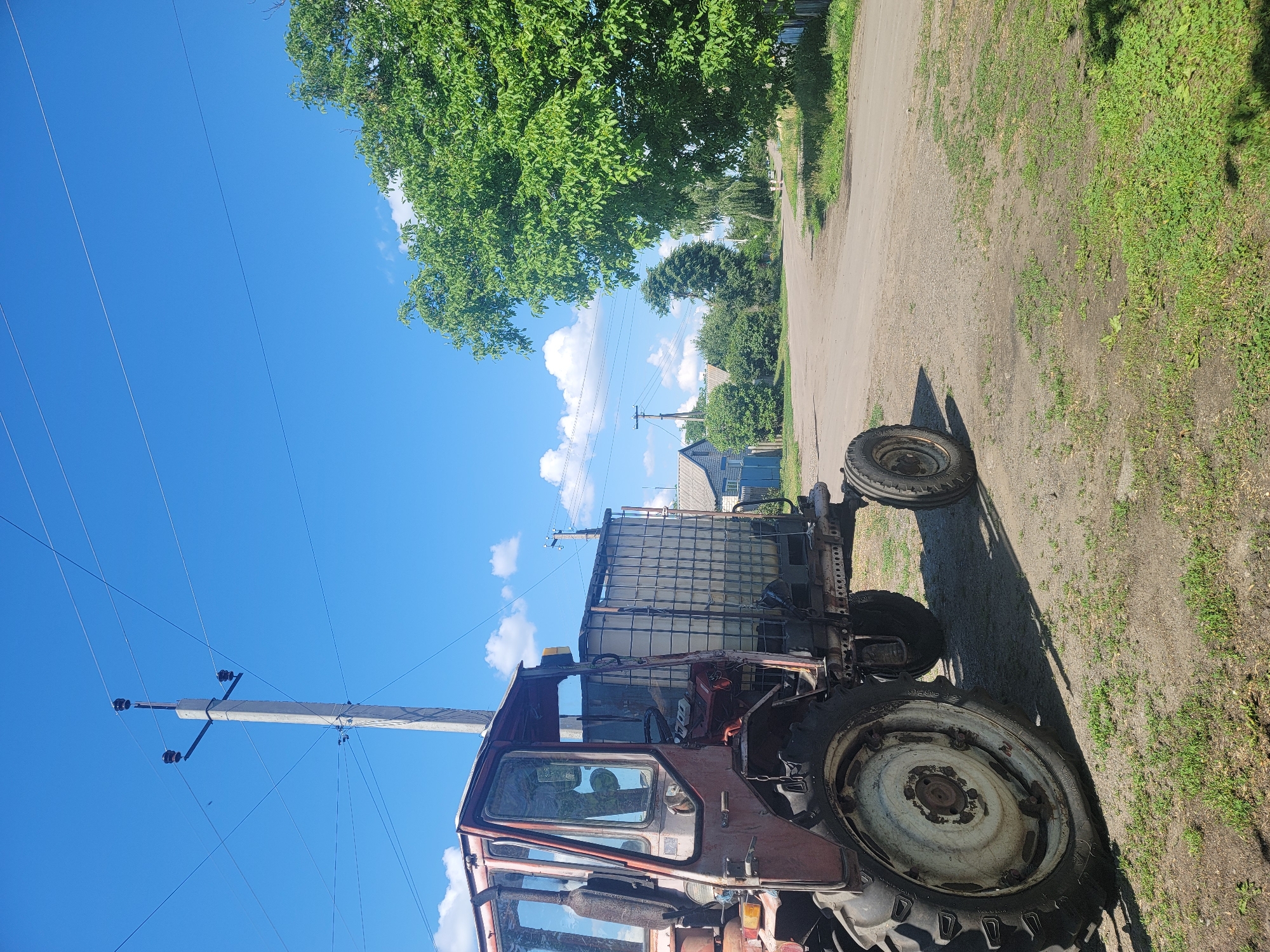 A low-angle, full shot captures a weathered, maroon tractor parked on a gravel road in Vovkivka, Ukraine. The tractor, which appears to be old and somewhat rusty, is equipped with a large, clear plastic water tank on its trailer. The tank is secured by a metal cage. The tractor's large rear tire is prominent in the foreground, casting a shadow on the ground.
Behind the tractor, a dirt road winds through a rural landscape. To the left, a concrete utility pole with wires stretches towards a bright blue sky dotted with fluffy white clouds. Further down the road, small, traditional houses with blue accents are visible amidst lush green trees and bushes. A large, leafy green tree dominates the right side of the frame, its branches reaching upwards. The overall impression is of a quiet, sun-drenched day in a rural Ukrainian village. There are no people visible in the image.