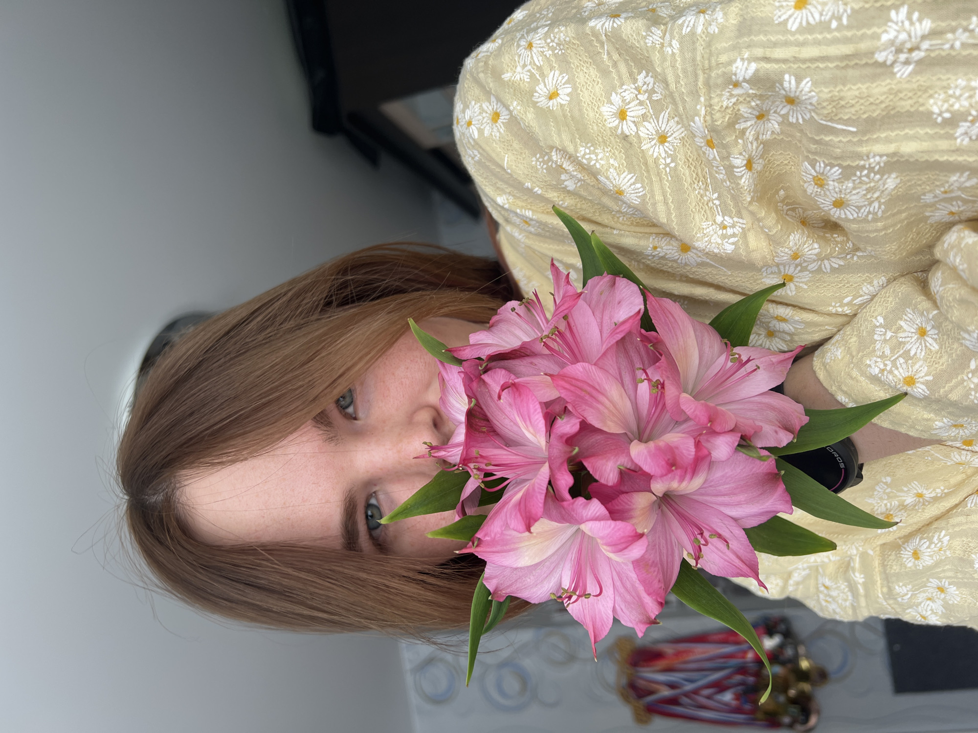The image presents a close-up portrait of an individual indoors in Chaikovsky, Russia. The person, appearing to be a young female, holds a bouquet of pink Alstroemeria flowers directly in front of their lower face, obscuring the nose and mouth. Visible features include light brown hair parted to the left, green-blue eyes looking directly forward, and freckled skin on the visible portions of the face. The individual is wearing a light yellow garment adorned with a pattern of white daisy-like flowers. A black smartwatch with white text displaying "GAROS" is visible on the left wrist, positioned below the flowers. The background features a wall with light blue and white abstract wallpaper on the left, beneath which a collection of medals with red, blue, and white ribbons is faintly visible. To the right, a dark, indistinct piece of furniture, possibly a cabinet or wardrobe, is present. The ceiling is light gray.