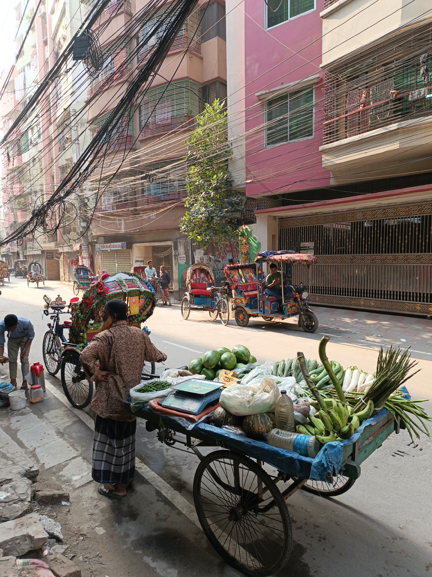 The image depicts a street scene in Dhaka, Bangladesh. Overhead, a dense network of electrical wires dominates the view. A vegetable vendor stands beside a three-wheeled cart laden with produce, including watermelons, green beans, bananas, and other vegetables, selling their goods on the side of a street. A customer in light-colored clothing is on the vendor’s right. In the background, several cycle rickshaws, one motorized rickshaw and people walk and sit along the street. Buildings line the street, one painted in pink.