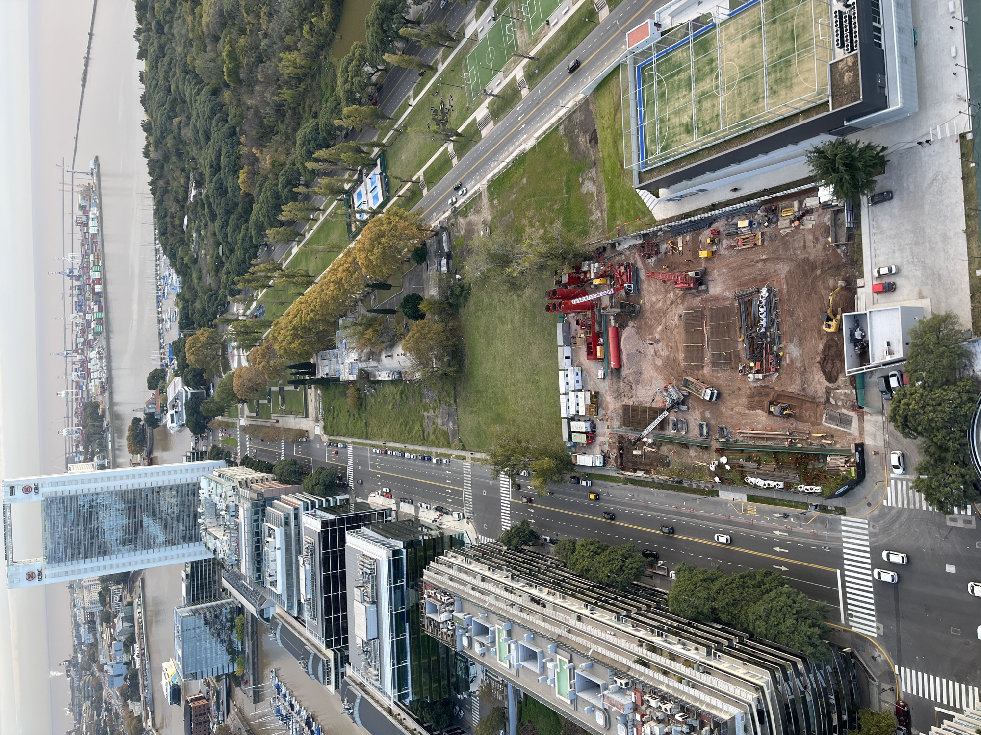 The image shows a high-angle view of a portion of Buenos Aires, Argentina. In the foreground, several modern high-rise buildings, likely apartments or offices, line a multi-lane road with visible traffic. The camera faces roughly west, with the Rio de la Plata and container port to the distant left. The Costanera Norte borders the river and a tree-lined paseo. A large construction site is adjacent to the buildings, with heavy machinery, including cranes and earthmovers, scattered around the site. A large recreational sports complex with several athletic fields can be seen in the upper-right corner of the image.