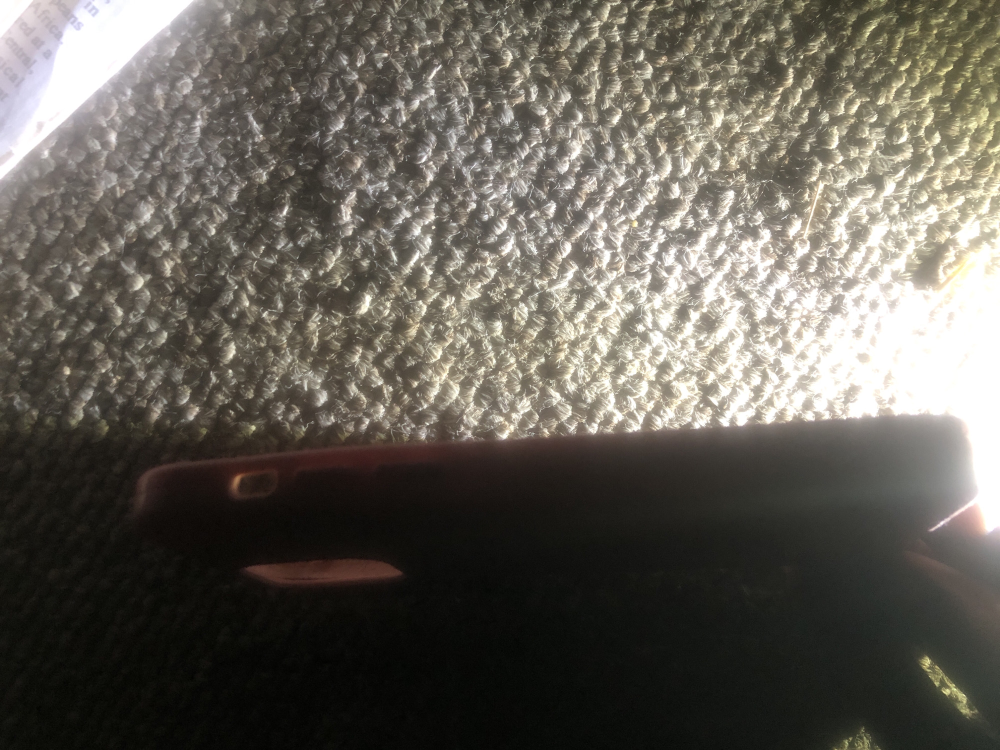 The image shows a close-up, angled view of a dark-colored smartphone case lying on a textured, brown carpet. The lighting is uneven, with a bright light source on the right side of the image creating strong highlights and shadows. The phone case appears to be red or dark pink. Part of the phone's side and a camera cutout are visible. The carpet has a looped pile texture. There is no discernible context related to the city of Jalingo, Nigeria, or any specific location within it. The image captures a still object in a dimly lit environment.