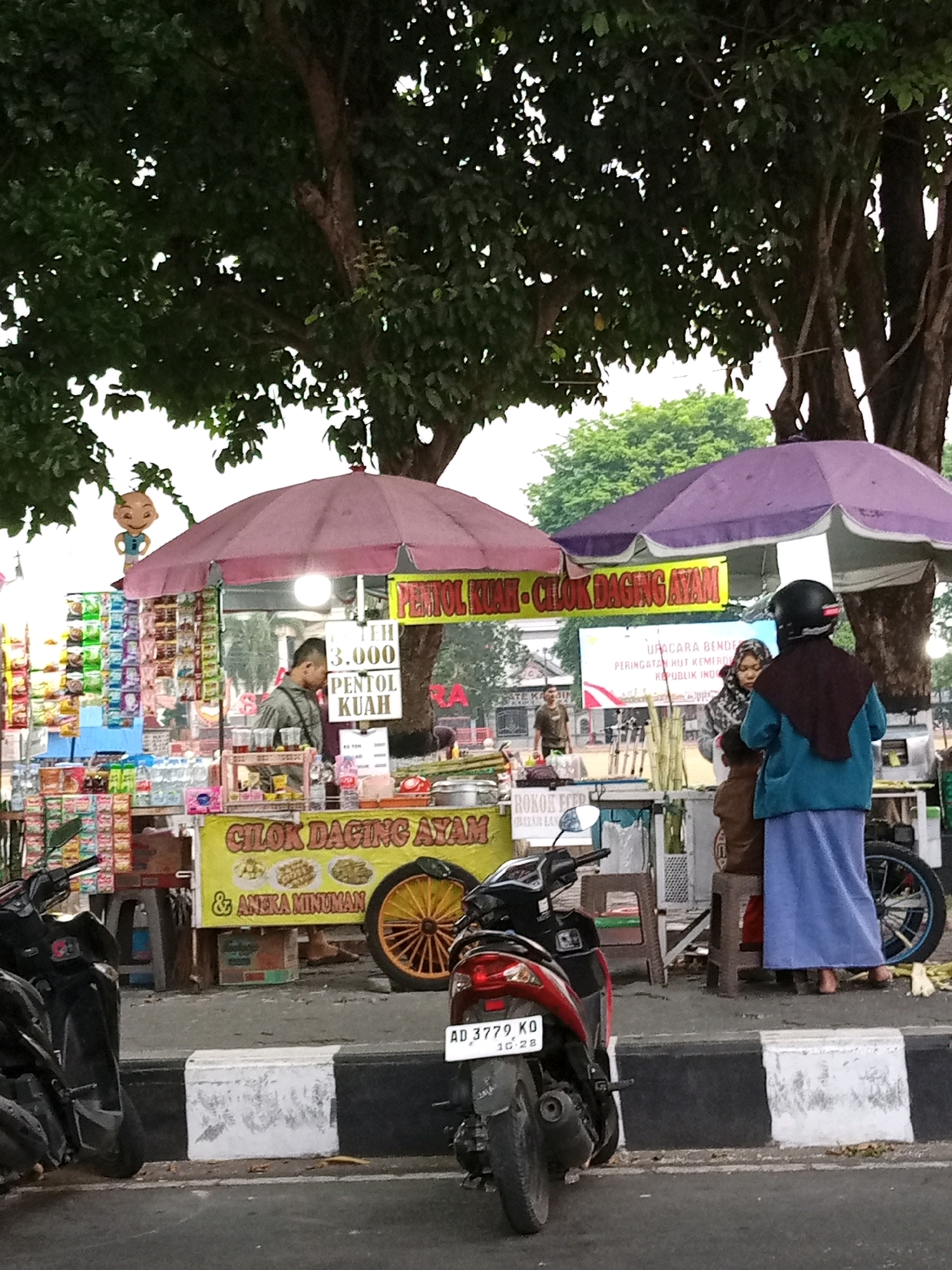 This is a ground-level, outdoor, eye-level shot of a street scene in Sukoharjo, Indonesia. The focus is on a food stall with two prominent umbrellas, one purple and one pink. The stall displays various food items and snacks, with colorful packaging stacked on shelves. A man is visible behind the counter, appearing to serve customers. Two women are interacting near the stall, one wearing a blue hijab and a turquoise top, the other in a patterned headscarf and brown clothing. A red motorcycle is parked in the foreground, with its license plate visible, reading "AD 3779 KO". The background features lush green trees, suggesting a somewhat natural or park-like setting within the city. The overall impression is of a vibrant street food vendor environment.