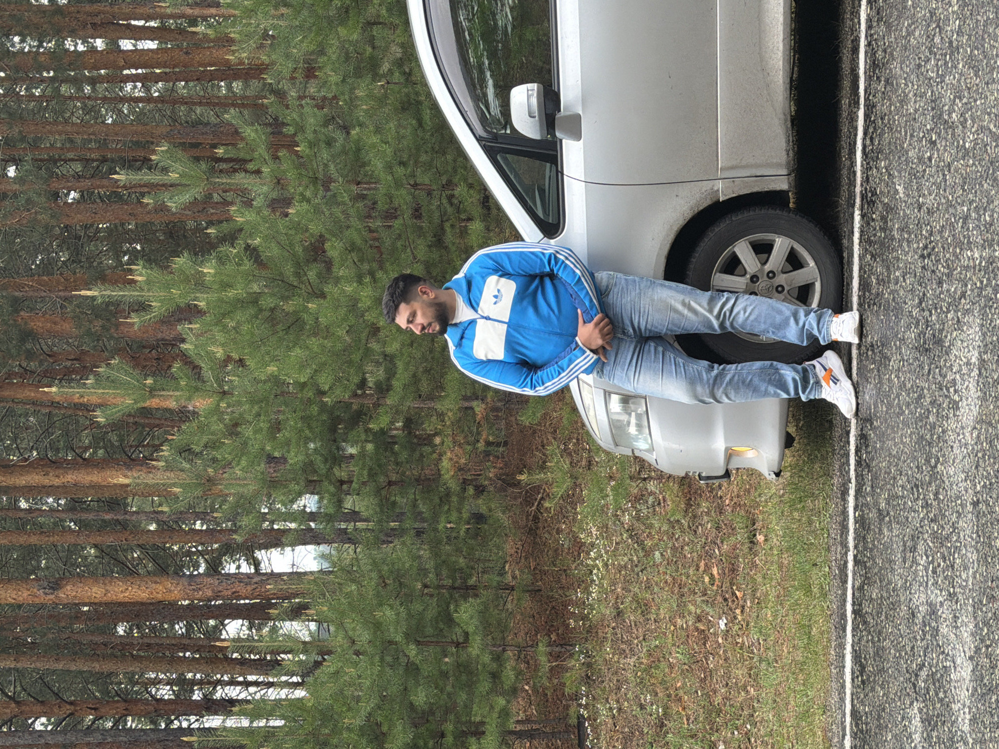 A man is leaning against a silver car parked alongside a road next to a forest. The man is wearing a blue Adidas track jacket, light-wash jeans, and white sneakers with orange stripes. The car is parked very close to the edge of the road. The road is paved and has a white line marking the edge. The forest consists of tall pine trees. The photograph was taken in Сизая, Russia.