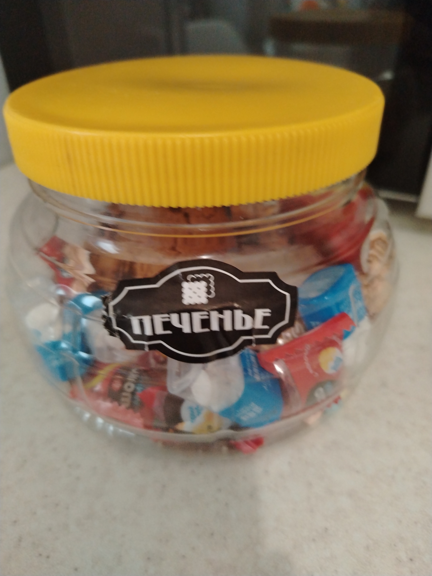 A transparent plastic jar filled with candies is on a speckled, light-colored surface, likely a countertop. The jar has a bright yellow, ribbed plastic lid. A black sticker with a white border and a white patterned graphic is adhered to the front of the jar. The sticker has white Cyrillic text that reads "ПЕЧЕНЬЕ" which translates to "COOKIES". Various individually wrapped candies in red, blue, and silver wrappers are visible inside the jar. The background is dark and out of focus, suggesting an indoor environment.