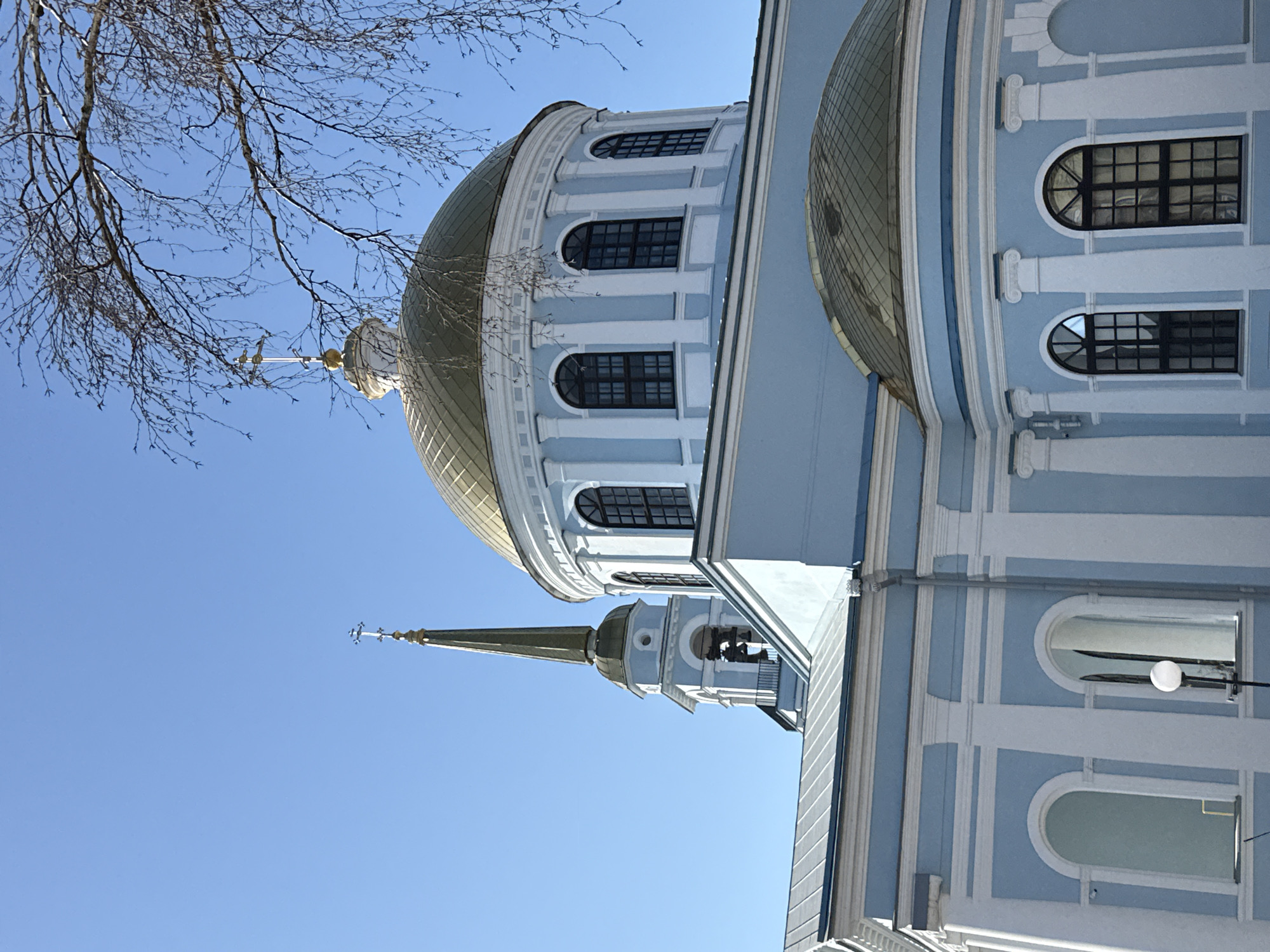 The image shows a building with a blue and white facade and golden domes, likely a Russian Orthodox church. The architecture features rounded windows with black frames, decorative columns, and a spire with a cross on top. A leafless tree is visible in the upper left corner. The sky is clear and blue. The location is likely Votkinsk, Russia, based on the architectural style of the building which is typical for Russian Orthodox churches.