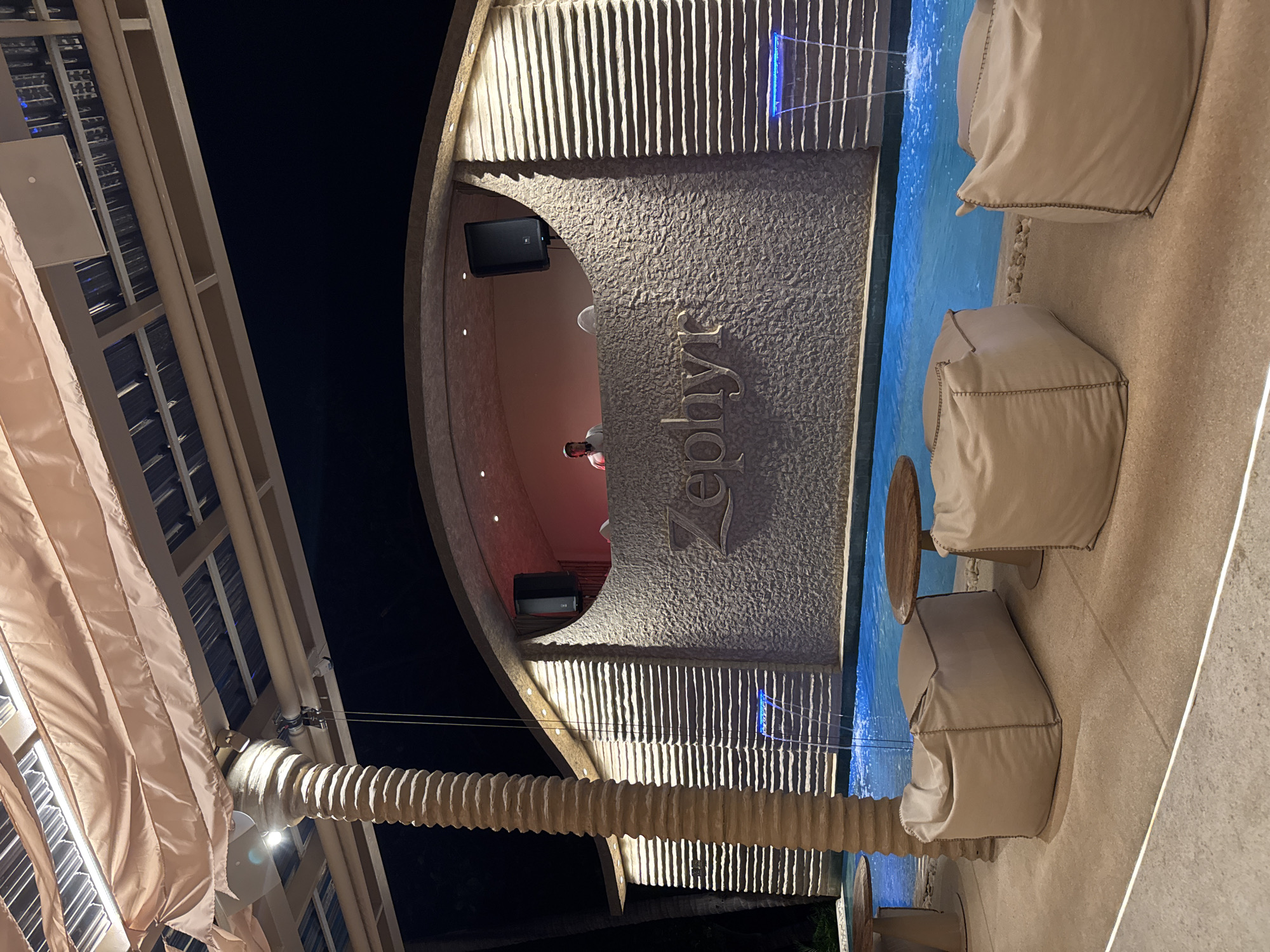 The image shows an outdoor lounge area at night, likely a bar or club, in Baan Lamai, Thailand. In the background, a DJ is visible behind a console with two speakers mounted above. The DJ booth is set against a textured, light-colored wall with the word "Zephyt" inscribed in large letters. The wall has horizontal grooves and two small waterfalls with blue lighting cascade into a pool in front of it. The pool is illuminated with blue light. In the foreground, there are three beige beanbag chairs arranged on a stone patio. Two small, round wooden tables are also present. A large, textured cylindrical column, possibly a decorative palm tree trunk, stands to the left of the frame. The area is covered by a translucent, peach-colored fabric canopy supported by a metal structure. The overall scene suggests a relaxed, evening ambiance with ambient lighting.