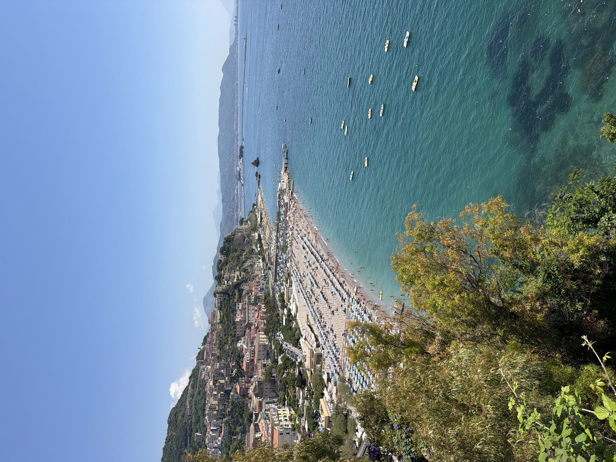 This aerial shot depicts a beach crowded with people and umbrellas in Vietri sul Mare, Italy, situated along a hillside dotted with buildings. The Tyrrhenian Sea stretches out to the right, dotted with small boats. Lush green trees and foliage frame the foreground. The sky is clear blue.