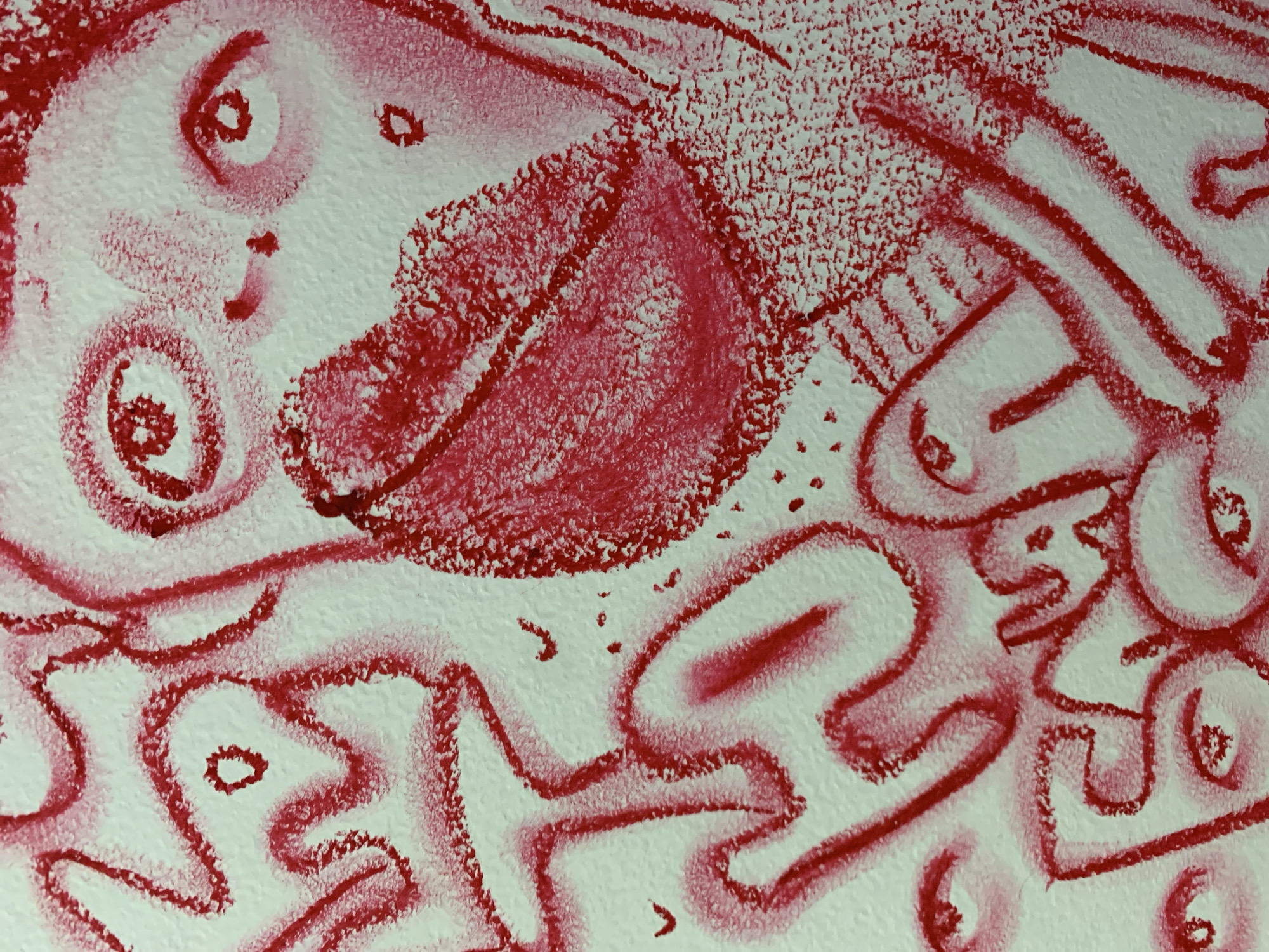 The image is a close-up photograph of a red crayon drawing on a white textured surface, possibly a wall. The drawing depicts abstract figures and facial features, including eyes, a nose, and a large mouth with prominent lips. The style is somewhat chaotic and expressive, with bold lines and areas of smudging. There are no discernible landmarks or elements within the image that would indicate its specific location in Kirov, Russia, or any other city. The photograph captures only the artwork itself, with no context of its surroundings.