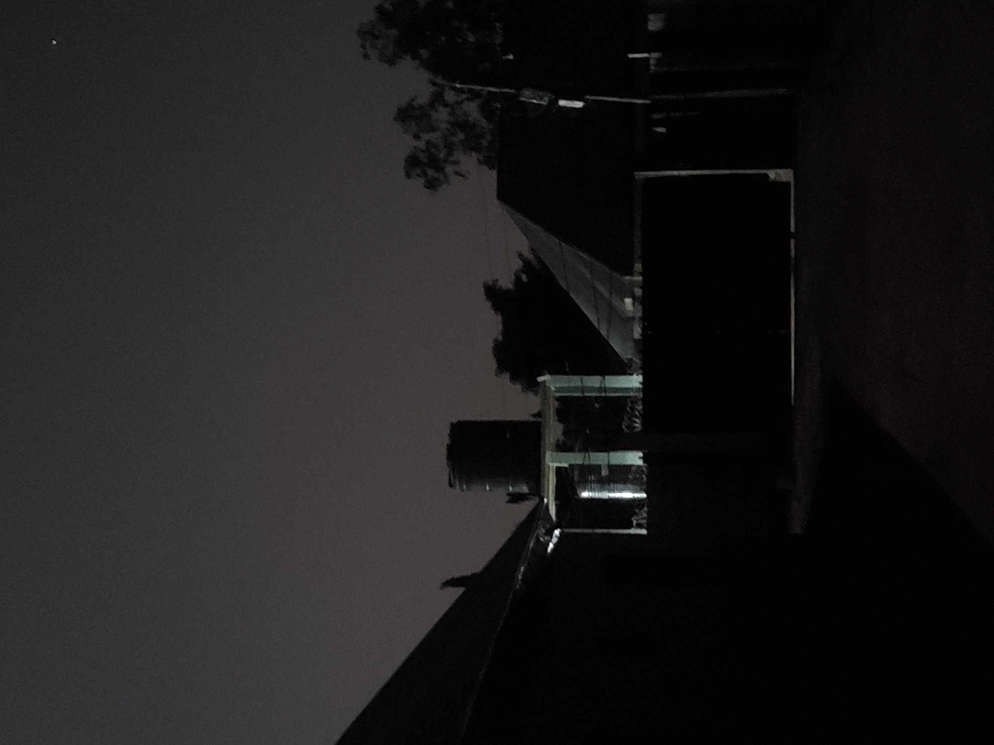 The image depicts a nighttime scene in Kason, Nigeria. In the foreground, a dark, angled structure, likely a roof or wall of a building, dominates the left side of the frame. To the right of this, a large, cylindrical water tank stands atop a lighter structure, possibly a small building or platform. This tank appears to be a prominent object in the scene. Further to the right, a building with a distinctive sloped roof is visible. The background features trees silhouetted against the dark night sky. A faint light source, potentially a distant star or light, is visible in the upper right portion of the sky. The overall impression is one of a residential or industrial area at night, with functional infrastructure like water storage being central to the composition.