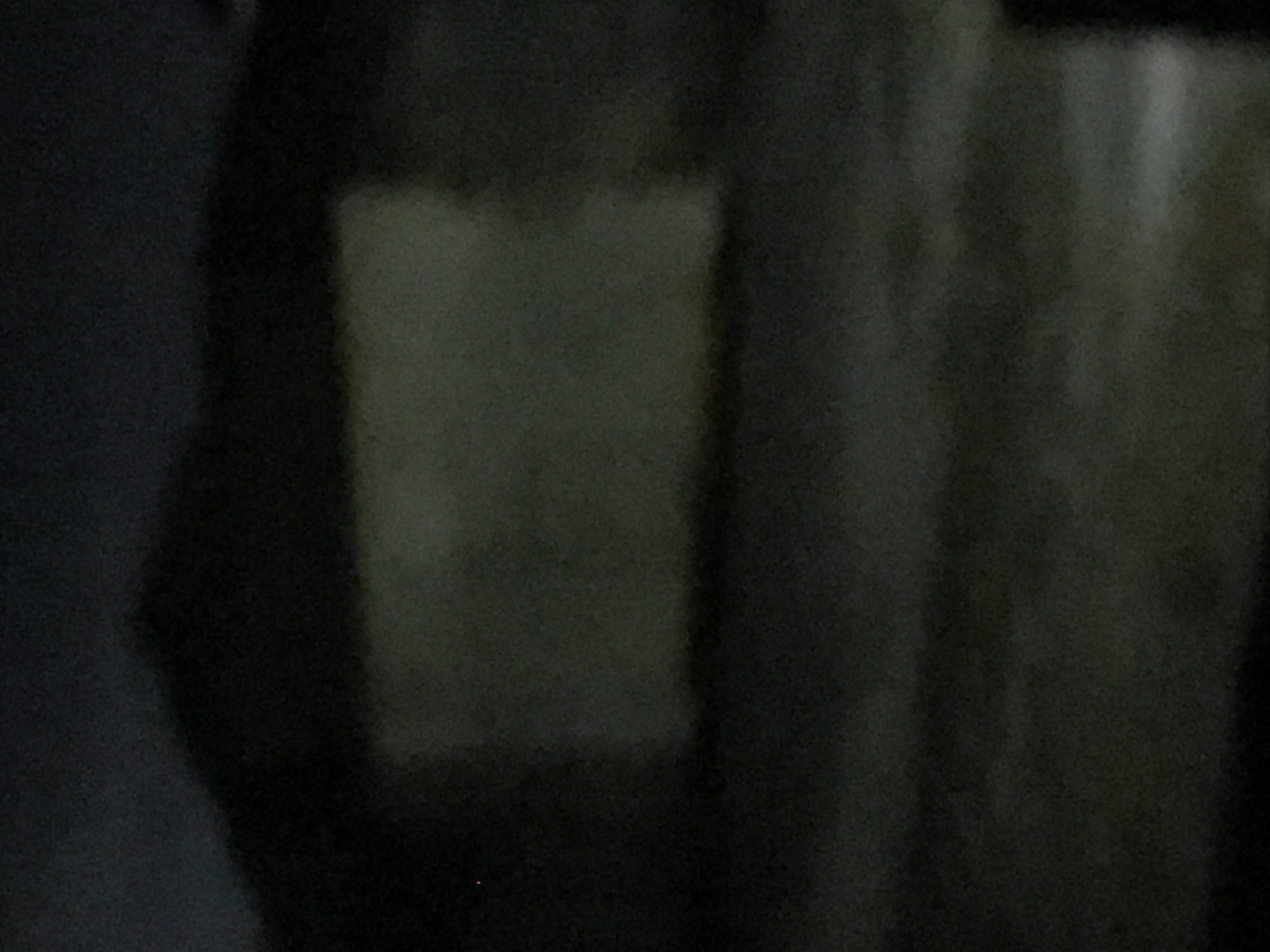 The image is a dark, blurry photograph of what appears to be a building. The building is largely obscured by darkness, with only a lighter rectangular section visible in the center, possibly a window or a lit portion of the structure. The foreground is also dark and indistinct, with no clear features. The overall impression is of an abandoned or derelict building at night. There are no people or discernible objects in the scene.