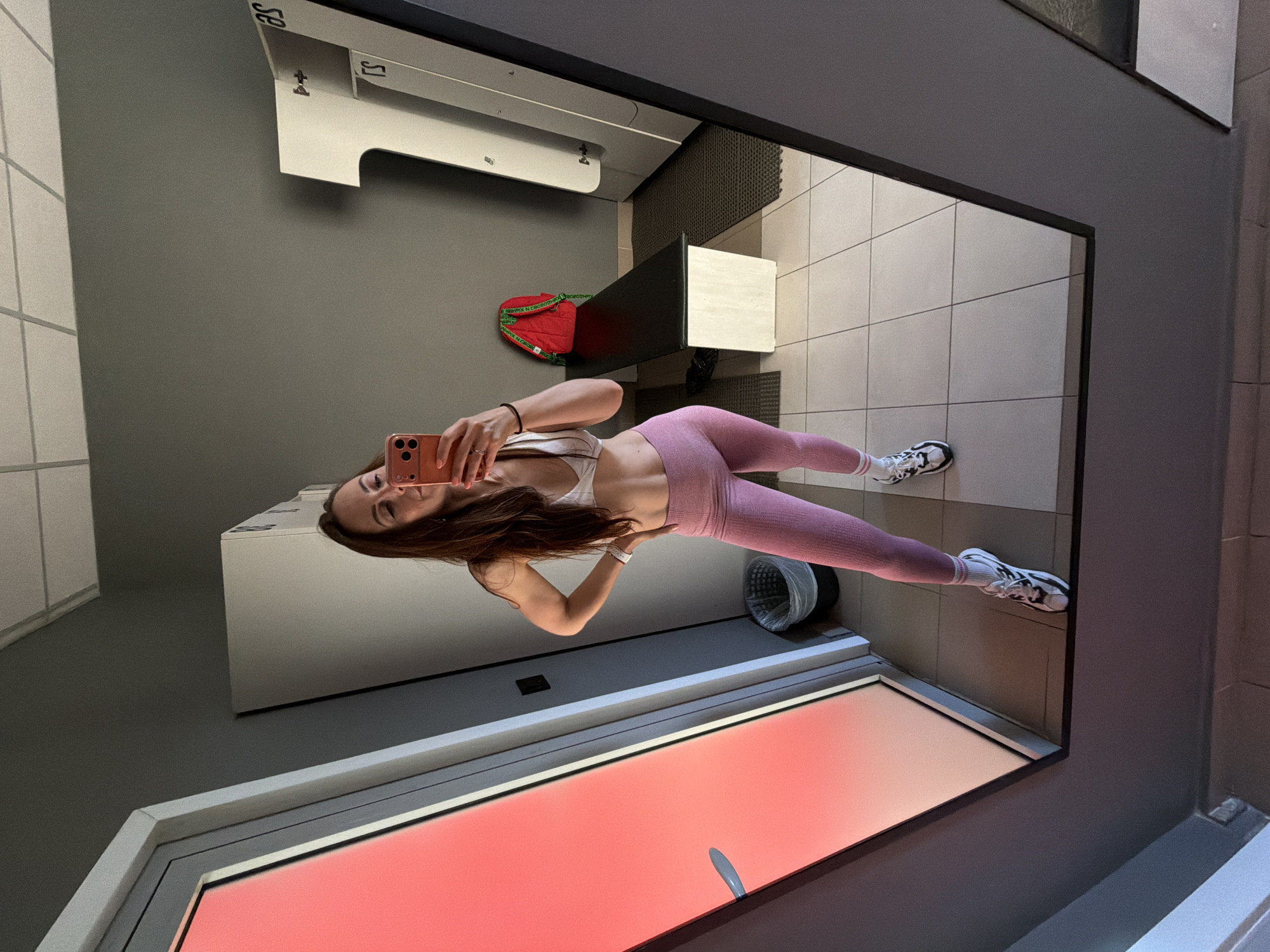 In a gym locker room, a woman stands by a large mirror, taking a selfie with her phone. She is wearing a white sports bra and pink leggings, with white and black athletic shoes. Her hair is long and brown, and she has a toned physique. The locker room has grey walls, tiled floors, and white lockers. A red backpack is on a black bench. To the left, a door with a red illuminated panel is visible. The image does not contain any information or clues that would indicate the location as Birobidzhan, Russia.
