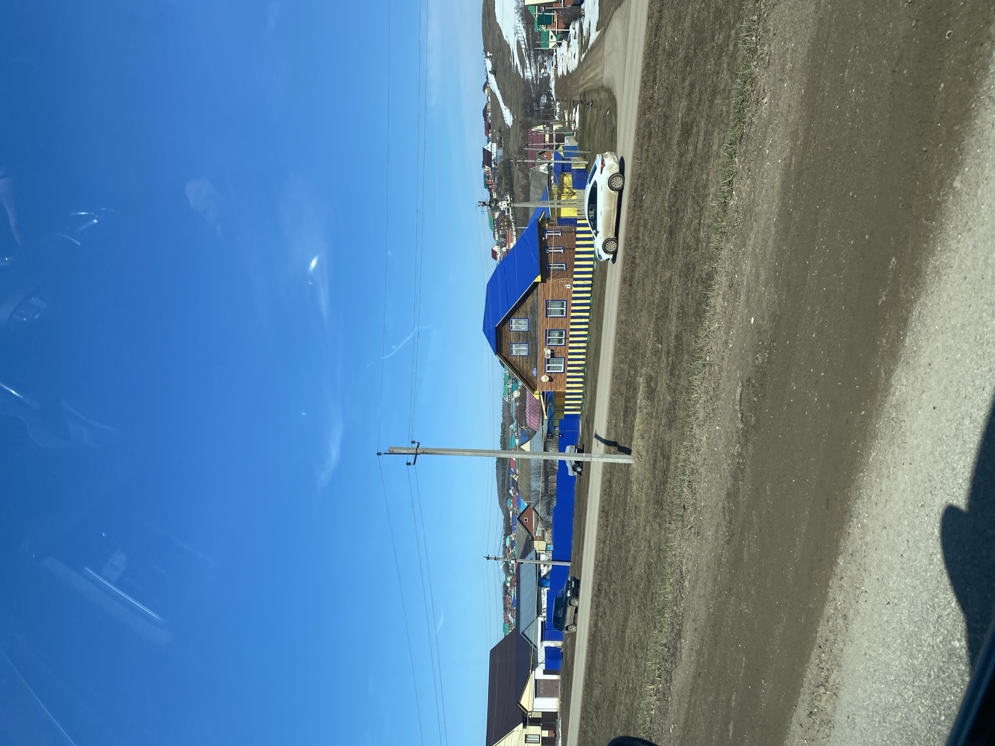 The image shows a street in Verkhniye Tatyshly, Russia. The view is from a moving vehicle. A bright blue sky occupies the left portion of the frame. On the right side of the road, there is a house with a distinctive blue roof and yellow and blue vertical stripes along its sides. A white car is parked adjacent to the house. Further up the road, a village or small town landscape extends into the distance. Utility poles and power lines run alongside the road. Patches of snow are visible on the distant hills. The roadside is earthen and grassy. The road itself is asphalt.